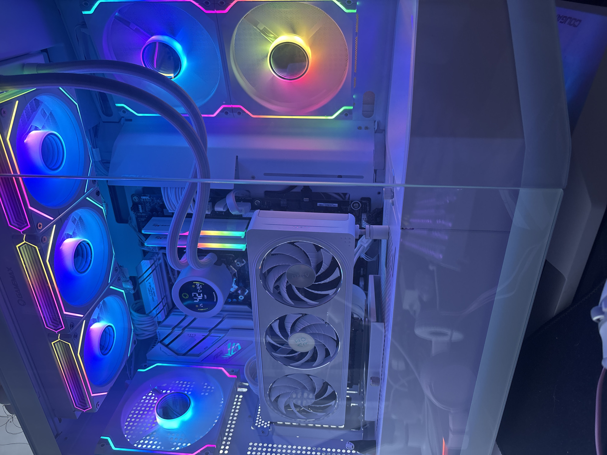 This is a close-up, eye-level shot of the interior of a white computer tower with a clear glass side panel. The computer is illuminated by vibrant RGB lighting, casting a colorful glow on the components. The top of the case features a white radiator with two large blue RGB fans. To the left, a large white case fan with diffused RGB lighting is visible. To the right, another large white case fan with a multi-colored RGB glow is present. A white graphics card with three white fans is mounted horizontally in the center of the case. A small circular LCD display, showing a temperature reading of 42 degrees Celsius, is attached to a white CPU cooler. White braided cables are neatly organized throughout the build. The bottom of the case is a white shroud, partially obscuring the power supply. The computer appears to be sitting on a dark surface.