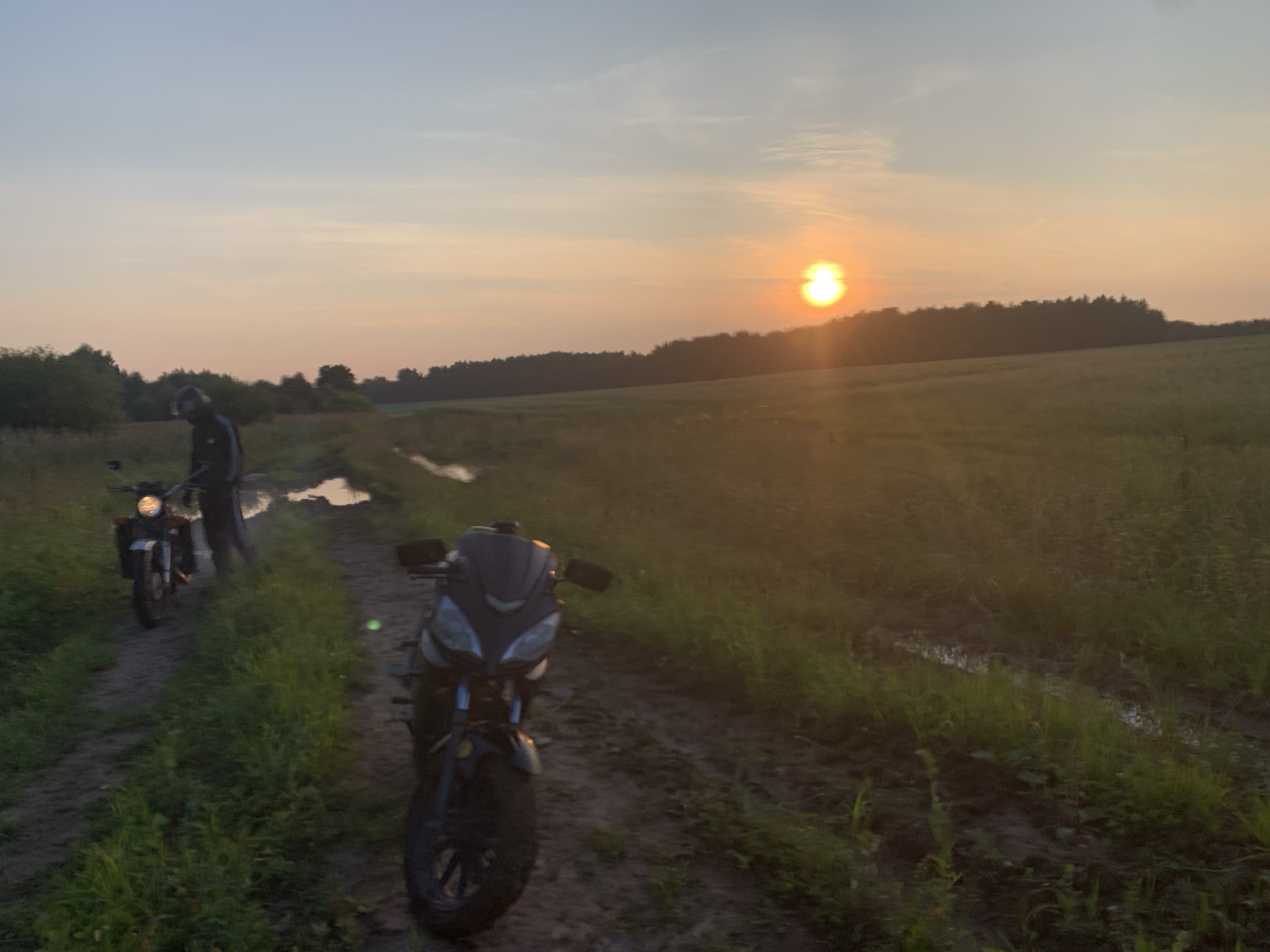The image depicts two motorcycles parked on a dirt track in a rural setting during sunset. The sky is a gradient of soft blues and oranges, with the sun low on the horizon, casting a warm glow. The track is muddy and has puddles of water, suggesting recent rain. Lush green grass and vegetation flank the track, and a line of trees forms the distant background.
In the foreground, a dark-colored motorcycle with a sleek design is parked, facing towards the right of the frame. Its headlights are off, and the front wheel is turned slightly. Further back on the left, another motorcycle, lighter in color and possibly an adventure bike, is parked. A person wearing a helmet and dark clothing is standing next to this motorcycle, appearing to be taking a break.
The overall atmosphere is peaceful and serene, capturing a moment of pause during a motorcycle ride in the countryside of Verkhnyaya Osh'ya, Russia. The lighting and natural elements suggest a natural landscape devoid of urban infrastructure.