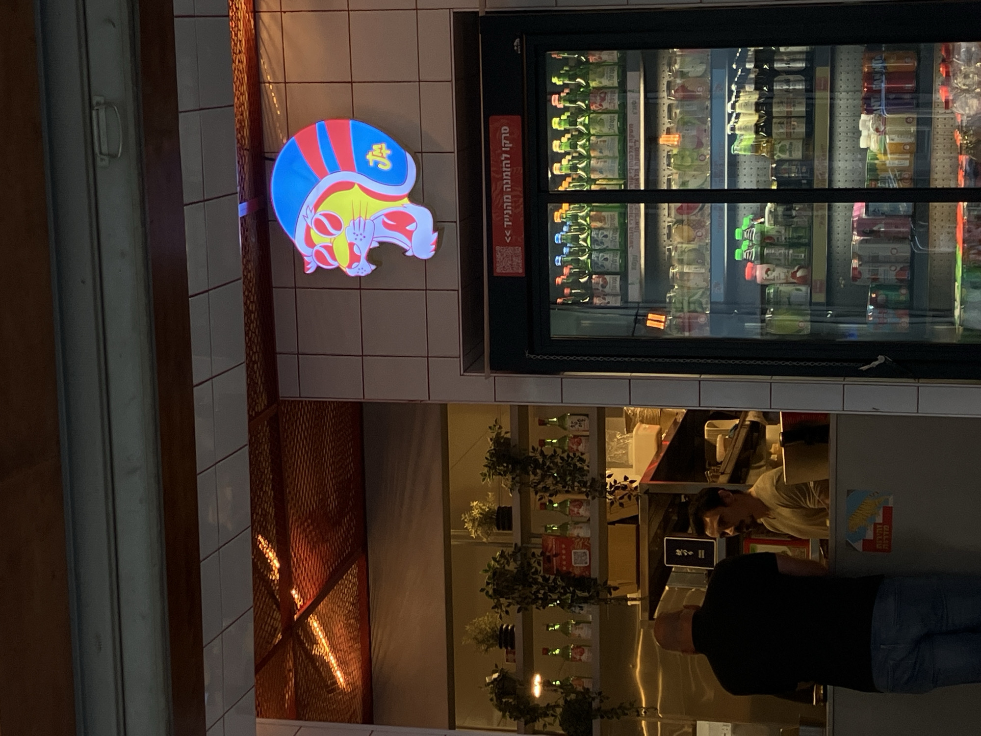 Here is a detailed description of the image:
**General Scene:**
The image shows the front of a business establishment, likely a convenience store or small eatery, at night. The scene is brightly lit, with the focus on the store's exterior and the activity inside.
**Location:**
Given the Hebrew signage and general aesthetic, the location is likely in Tel Aviv, Israel.
**People:**
Two people are visible inside the establishment:
1. **Person 1:** Wearing a black shirt and jeans, facing the camera. It is unclear if they are a customer or an employee.
2. **Person 2:** Partially visible behind a counter, possibly an employee, looking towards Person 1. They are wearing a light-colored shirt.
**Subjects & Objects:**
* **Storefront:** The store has a tiled facade with a distinctive, colorful neon sign of a cartoon-like character above the entrance.
* **Refrigerated Display:** A glass-fronted refrigerator unit is visible, filled with various beverages and food items. There is Hebrew writing on a vertical sign attached to the fridge.
* **Interior:** Shelves decorated with greenery.
* **Counter Area:** A counter area is visible with various items and equipment, suggesting a service area.
**Actions & Interactions:**
* The two people visible inside are likely interacting, although the specific nature of their interaction is unclear. One person appears to be observing the customer, while the customer's actions are not clearly discernible.
**Environment:**
The scene is an urban environment at night. The lighting is bright, illuminating the storefront. The overall impression is of an active, bustling street scene.