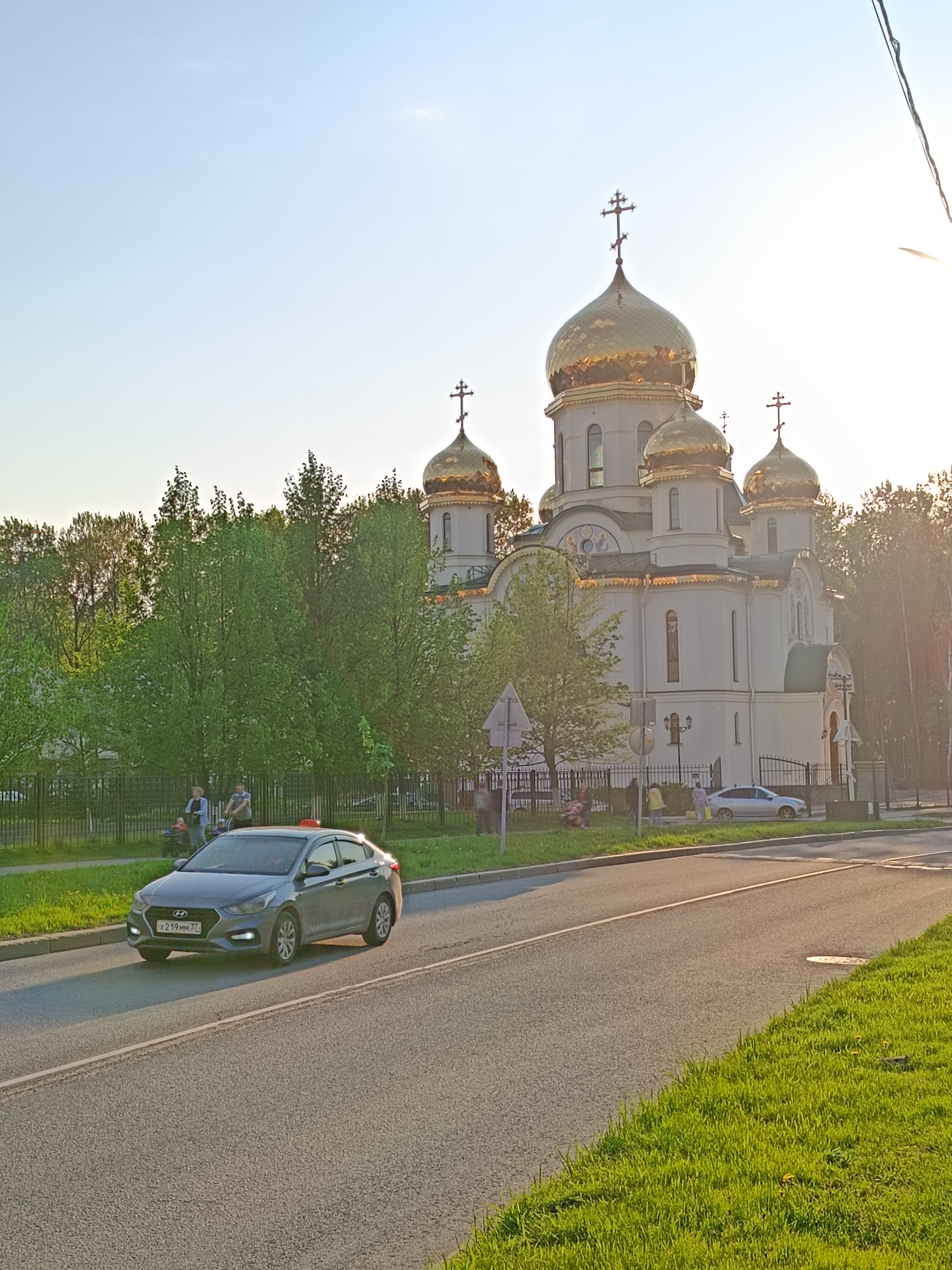 Captured on a bright day in Saint Petersburg, Russia, the scene features a light grey Hyundai car with the license plate "Х219ММ 37" traveling on an asphalt road. To the right, a grassy patch extends towards the street. On the opposite side, a group of people, including individuals with a stroller, is positioned near a fence line and green trees. A white Eastern Orthodox church with golden domes rises prominently in the background. A traffic sign is visible near the church. Sunlight is present on the right side of the frame.