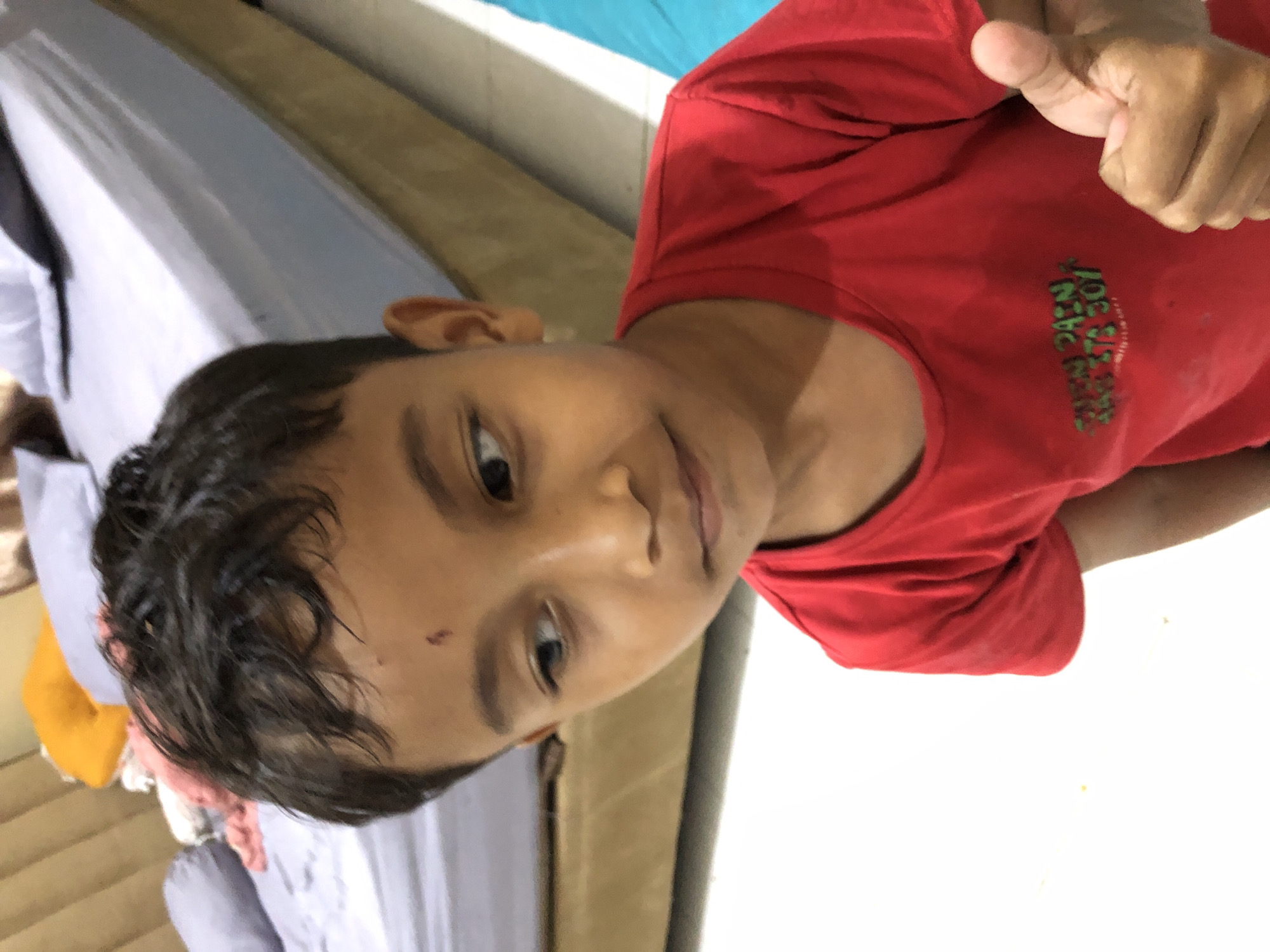 A young boy with dark hair and brown skin is looking to the left, with a small red mark on his forehead. He is wearing a red t-shirt with a graphic design on it. His right thumb is raised in a thumbs-up gesture. The background shows a bed with grey and purple bedding, and a sliver of a blue object. The scene appears to be indoors.