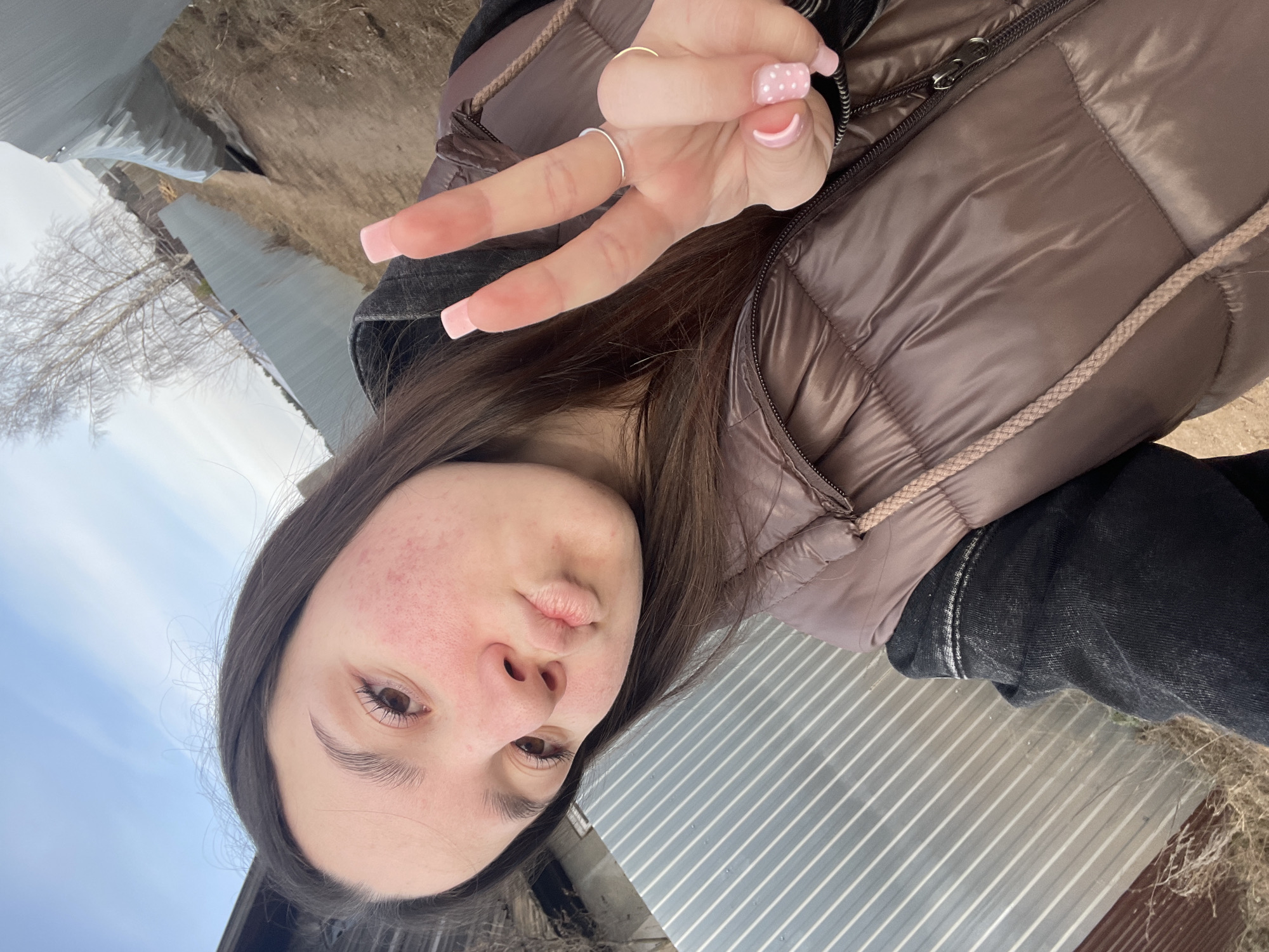 A young woman is taking a selfie outdoors in what appears to be a rural or semi-rural area. She is wearing a brown puffer vest over a dark colored hooded sweatshirt. She is making a peace sign with her left hand, which features light pink manicured nails; one has white polka dots. She is also wearing two rings on the same hand. The background includes a building with corrugated metal siding and a cloudy sky, suggesting an outdoor location. The specific location is Ольховка, Russia. Dry grass and bare trees are also visible in the background.