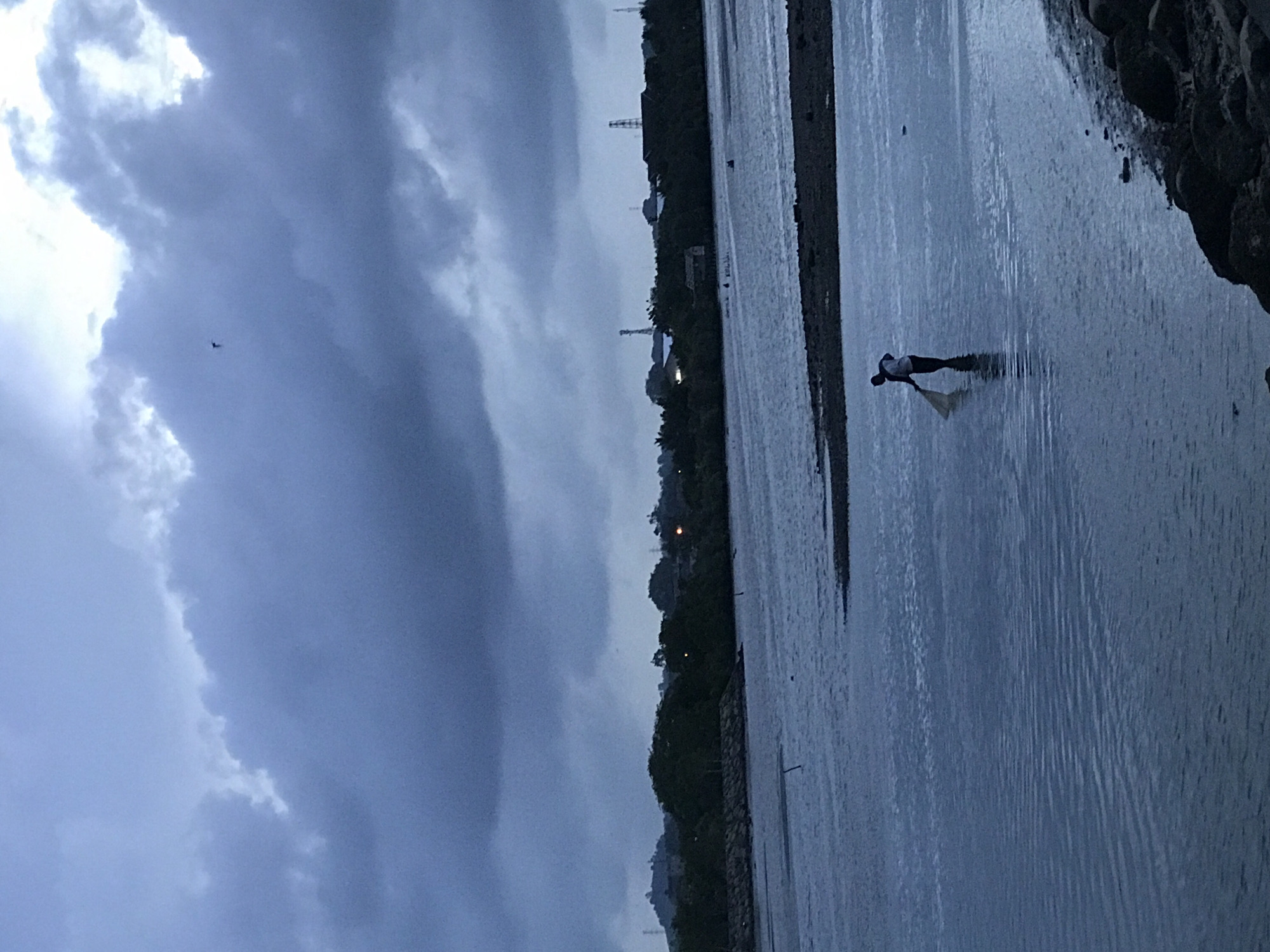 A wide-angle shot shows a silhouetted figure in a body of water under a cloudy sky. The person stands in shallow water near a shoreline, bending over with an object in their hands, possibly fishing or collecting something. The water's surface is textured, suggesting movement or current. The opposite bank is lined with trees and structures, some of which appear to be industrial based on the presence of towers. In the sky, a small, dark object resembling a plane is visible amidst the cloudy formations. The scene is likely near Lhokseumawe, Indonesia, though specific landmarks are not visible to verify the location. The overall atmosphere is muted and slightly overcast.