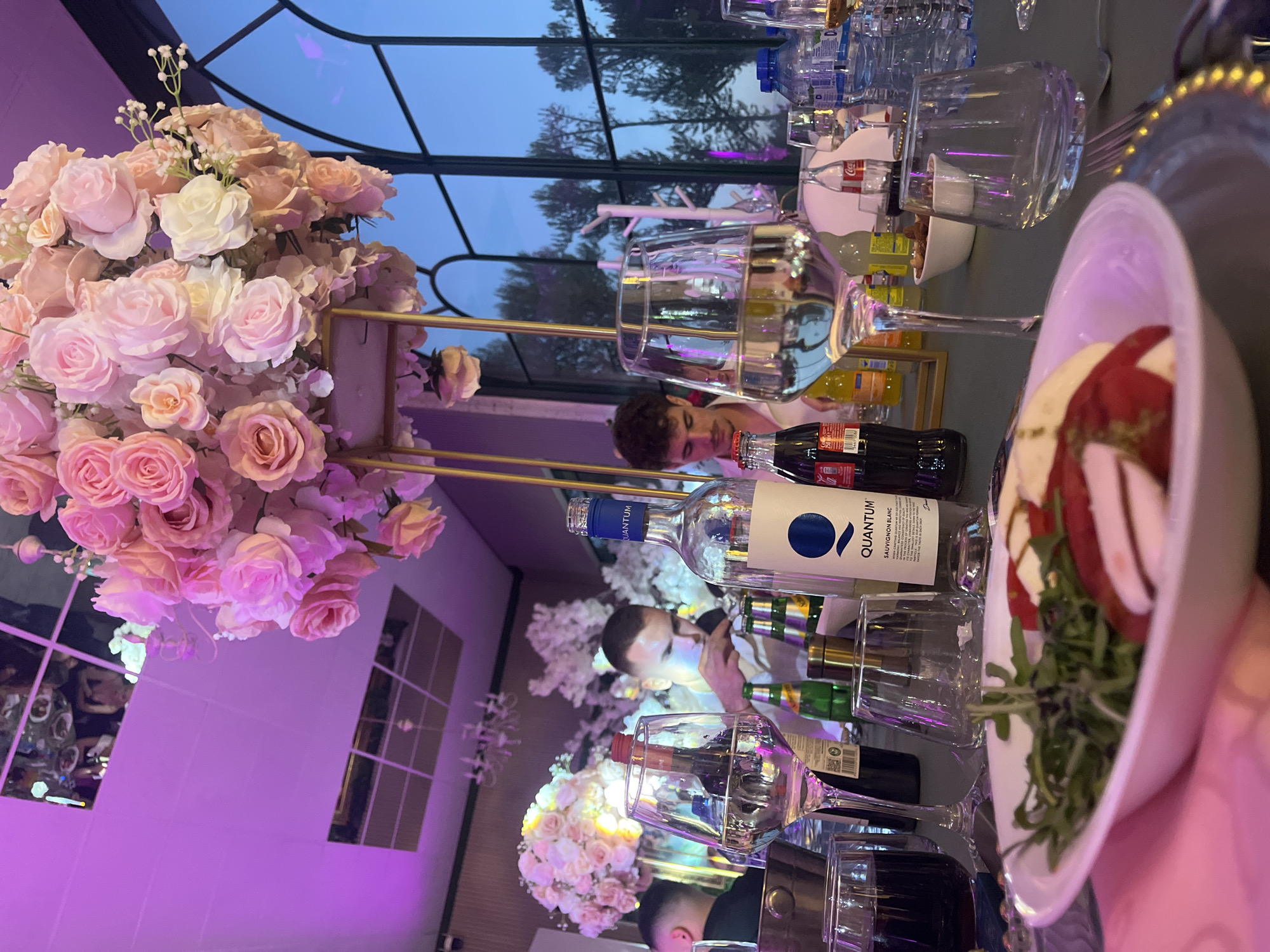 The image captures an elegantly decorated table at an indoor event, likely a dinner or celebration, situated in Plovdiv, Bulgaria. A prominent centerpiece features abundant pink and white roses along with baby's breath, arranged on a gold-colored stand. The table surface is reflective, creating intricate mirror images of the surroundings and guests.
Through large windows in the background, a twilight sky with silhouetted trees is visible, indicating that the time of day is dusk or early evening. The interior is illuminated with ambient purple and pink lighting, contributing to a festive atmosphere.
The table is set with various items including wine glasses, some containing beverages, and multiple water bottles. Several Coca-Cola bottles are visible, alongside a bottle of "Quantum" Sauvignon Blanc wine, which has its label clearly legible. A plate holding a dish resembling a caprese salad, with slices of tomato, mozzarella, and arugula, is positioned on the table.
Reflections of people are discernible in the mirrored table surface and through the glassware. One individual with dark, curly hair is partially visible, leaning over the table. Other indistinct figures are reflected in the background, suggesting multiple attendees at the gathering. The scene conveys a sense of a well-organized and pleasant social event.