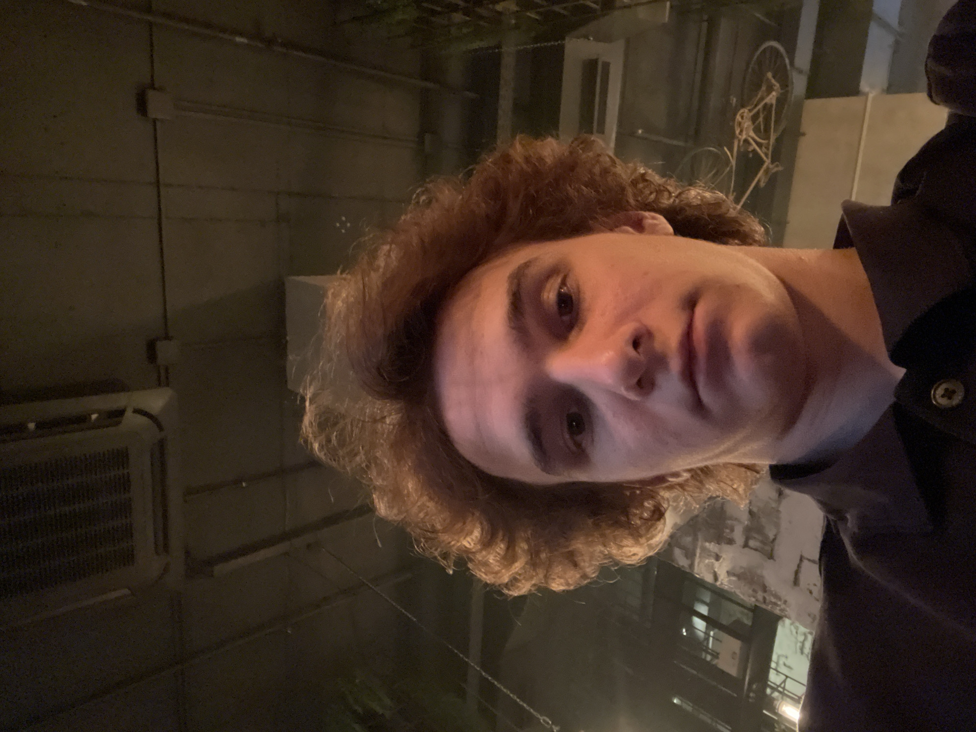 A low-angle, close-up selfie shows a young man with curly, light brown hair looking directly at the camera. He is wearing a dark collared shirt. The background consists of a dimly lit interior with exposed ceilings, ductwork, and some hanging chains. The overall impression is that of an industrial or modern commercial space, possibly a bar or restaurant. The specific location in Krasnoyarsk, Russia cannot be determined from the visual information provided in the image.