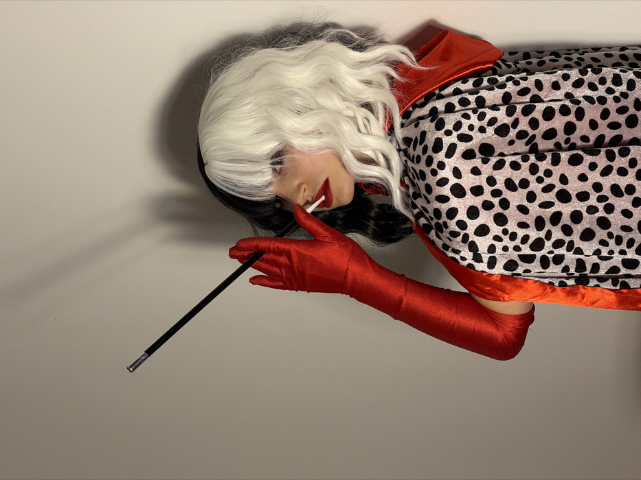 The image depicts a female individual in a half-body shot, presented in left profile, against a plain, light-colored wall background, likely indoors. The subject wears a distinctive costume. Her hair is a wig, split vertically down the middle, with black on the left and white, wavy hair on the right, featuring bangs across the forehead. Red lipstick is applied to her mouth. She is wearing long, shiny red gloves that extend past the elbows. A cape, featuring a black-spotted pattern on a light background (resembling a dalmatian print) with a visible red lining, covers her shoulders and back. In her gloved right hand, she holds a slender, black cigarette holder with a silver tip, which has a white-tipped object (likely a cigarette) inserted into its end, positioned at her mouth. Shadows are cast on the wall behind her, indicating a light source. The specific location within the city of Budva, Montenegro, cannot be determined from the visual information provided in the image.