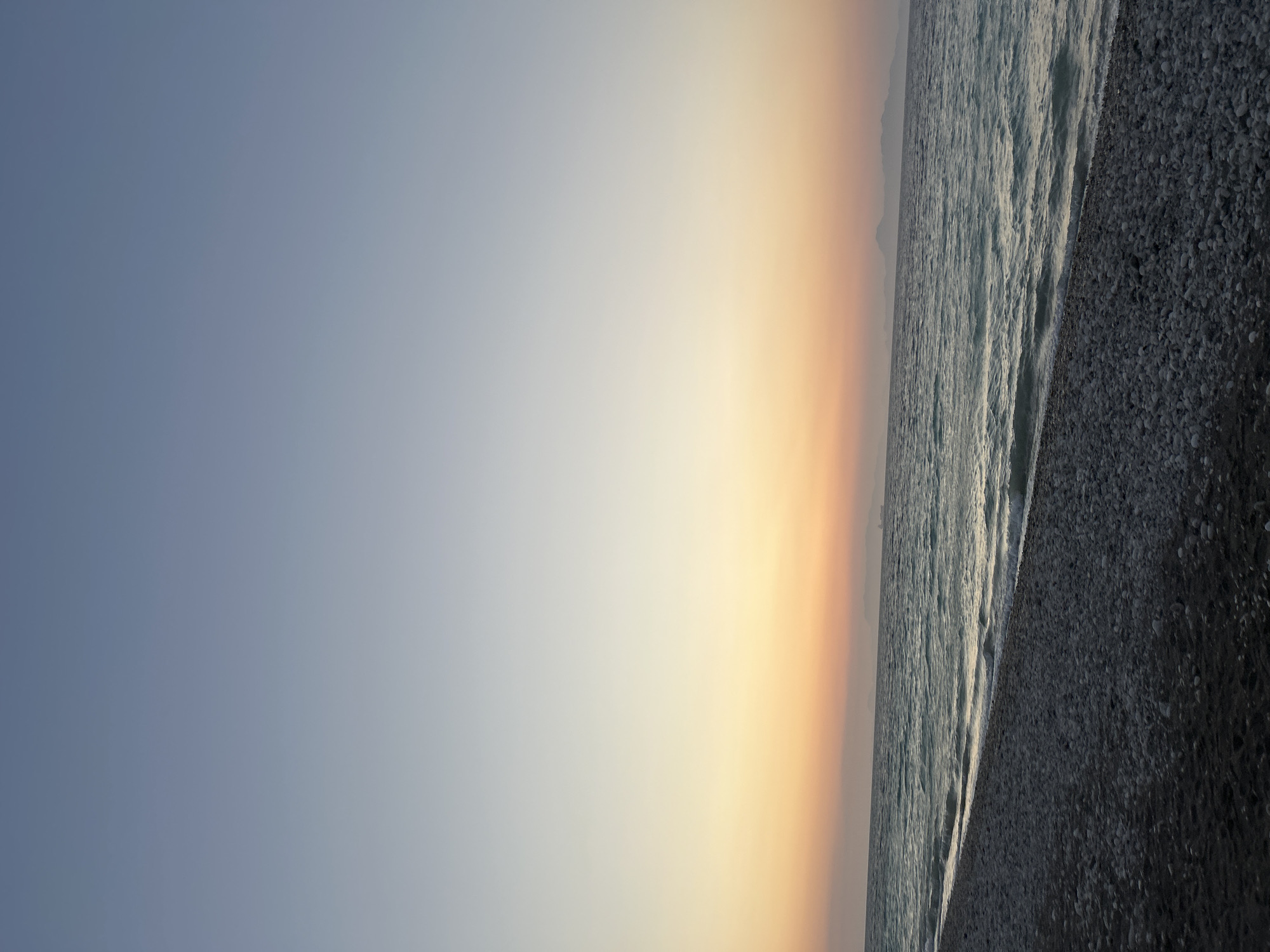 The image captures a serene coastal scene at what appears to be sunset or sunrise in Rhodes, Greece. The foreground is a dark, pebbled beach. The middle ground is occupied by the Aegean Sea, its surface rippled by gentle waves that break upon the shore. In the distance, a hazy silhouette of islands can be observed. The sky transitions from a pale blue at the top to a soft orange and pink hue near the horizon, suggesting the low angle of the sun. There are no people or man-made structures visible on the beach itself. A faint shape of a ship is visible on the horizon.