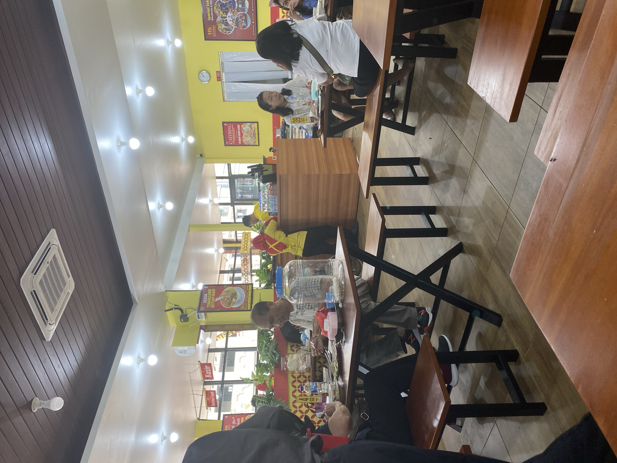 This is a photograph taken inside a casual dining restaurant in North Jakarta, Indonesia. The interior features a yellow-painted wall and a dark wood-paneled ceiling with recessed lighting and an air conditioning unit. The restaurant is furnished with simple wooden tables and benches.
In the foreground, a man is seated at a table, eating. He is wearing a white t-shirt and grey pants. In front of him are plates of food, cutlery, a bottle of water, and a large glass dispenser. To his left, another person is partially visible, also seated at the table.
In the middle ground, several other patrons are dining at separate tables. Two women are seated at a table, with one appearing to be speaking. Another woman is visible further back, also at a table. Behind the counter, there are two staff members wearing yellow uniforms. One is visible from the back, interacting with a customer, and another is standing behind the counter. Various signs are displayed on the walls, including advertisements for "Catering" and "Sedia Tengkleng Setiap Hari" (Ready Tengkleng Every Day).
The overall scene depicts a busy, everyday dining experience in a local eatery. The flooring is tiled with a light brown pattern. The lighting is bright, indicating it is daytime or the restaurant is well-lit.