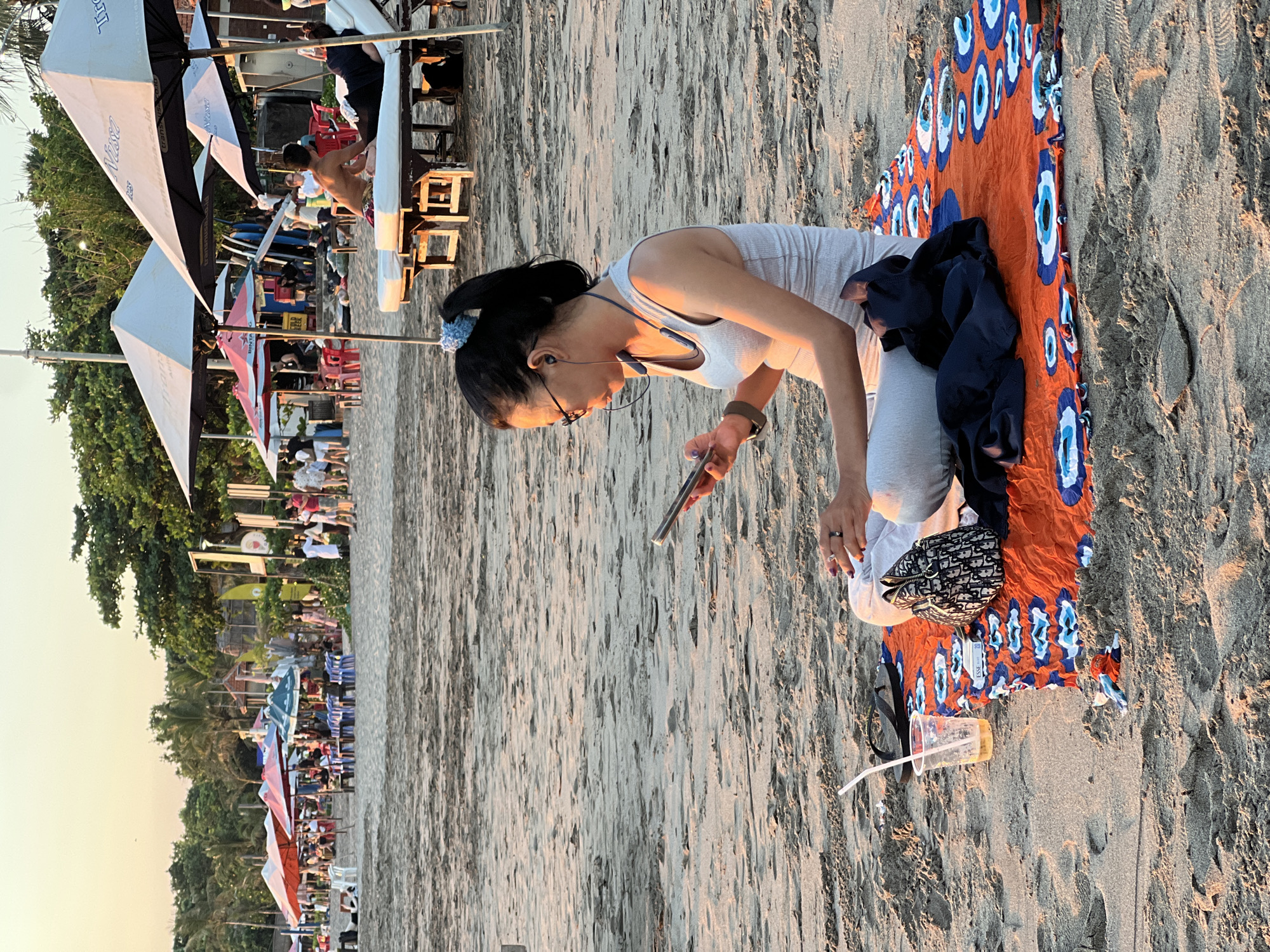 The image, captured on a sandy beach in Kuta, Indonesia, depicts a lively late afternoon or early evening scene. In the foreground, a woman with dark hair pulled back, wearing glasses, a light grey tank top, and light grey pants, is kneeling on the sand, engrossed in her smartphone. She wears a neckband headset, a watch on her left wrist, and a ring on her right hand. Her posture suggests focused attention on her device.
She is situated next to an orange and blue patterned sarong, adorned with a distinctive 'evil eye' motif. A dark jacket or shirt is draped on the sarong, alongside a small, black and white patterned handbag, whose design is reminiscent of a Dior saddle bag. Nearby, a plastic cup containing a drink and a white straw rests on the sand. The immediate sand appears textured with ripples and footprints.
The background reveals a bustling beach environment with numerous people. A row of beach umbrellas, some white and black bearing the text "Tiara" and partial website information, and others displaying "BINTANG" (a local beer brand), line the shore. Many individuals are visible, relaxing on beach chairs under the umbrellas, walking, or socializing. Lush green tropical foliage and trees form a dense band along the edge of the beach. The sky, visible in the upper part of the image, exhibits a clear, warm hue, indicative of the soft, golden light of sunset or late afternoon. The overall atmosphere conveys leisure and enjoyment at a popular coastal destination. A small tag with "ESS" is visible on the sarong.