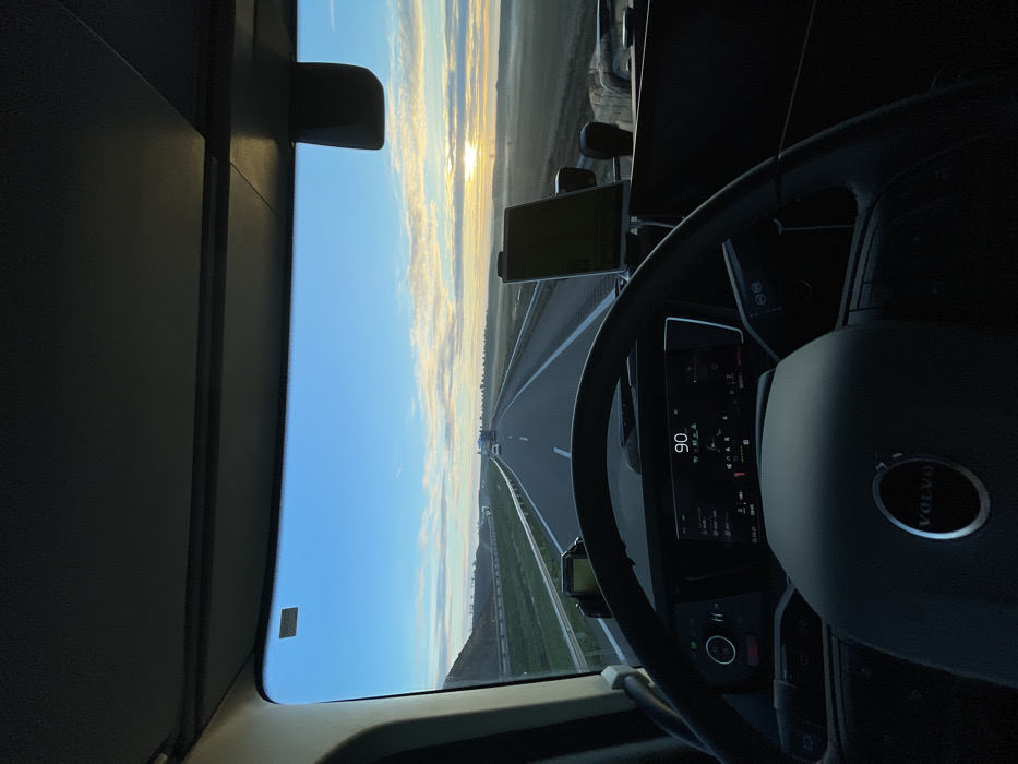 Inside a Volvo truck cab, the view faces forward onto a highway under a sunset sky. The dashboard displays a speed of 90. The steering wheel, partially obscuring the dash, features the Volvo logo. A device mounted on the dashboard is visible to the left. The outside world consists of a straight, multi-lane highway stretching into the distance, flanked by grassy fields. The sky transitions from blue to hues of orange and yellow near the horizon where the sun is setting.
