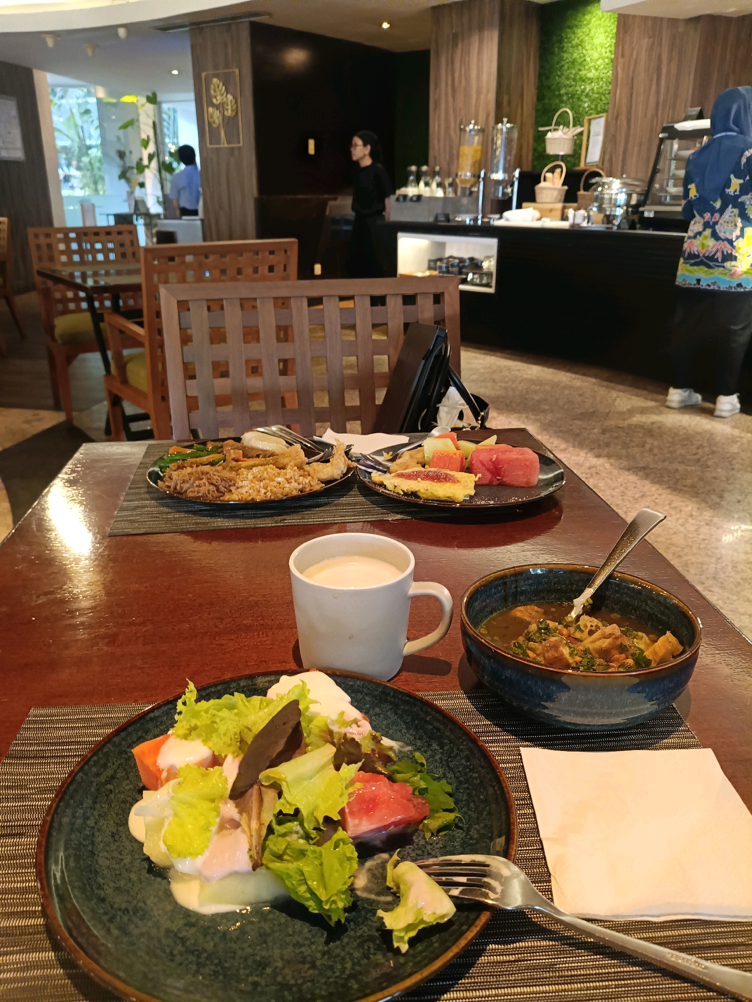 The image depicts a breakfast buffet setting at a hotel, likely in North Jakarta, Indonesia. In the foreground, a wooden table is set with plates of food and drinks. A dark green plate holds a salad with lettuce, what appears to be watermelon and papaya, and a creamy dressing. Next to it, a deep blue bowl contains a hearty stew with meat and vegetables, with a spoon resting inside. A white mug, likely containing coffee or tea, is also on the table, as is a white napkin. Further back, a plate of fried chicken and other savory items is visible, along with a plate of mixed fruits. The background reveals a buffet station with various food dispensers and serving dishes. In the distance, several people are present, including a woman in a black dress and another in a blue headscarf and patterned clothing. The overall ambiance suggests a casual dining experience in a hotel restaurant.