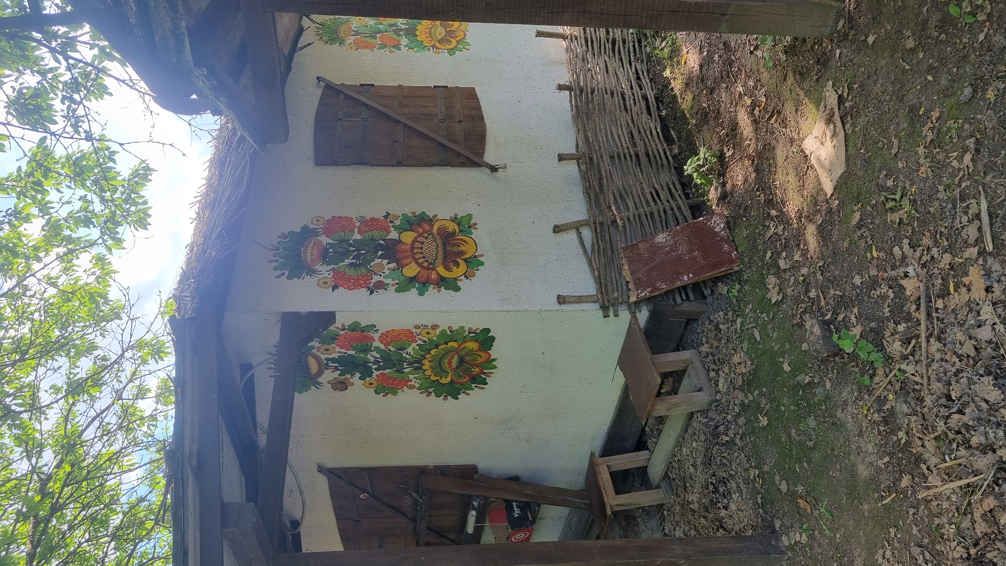 The image depicts the exterior of a traditional, single-story building in what is identified as the city of Buda, Ukraine. The structure features whitewashed or light-colored stucco walls and a thatched roof, partially covered by wooden beams. The visible wall is adorned with two large, vibrant folk art floral patterns. The left pattern prominently displays red berries and green foliage, with a central large yellow and orange flower. The right pattern mirrors this style, also featuring red berries, green leaves, and a large central yellow and orange flower.
On the left side of the building, an open wooden entry reveals a dark interior containing shelves with various objects, including a rectangular item with the visible text "Poguuy." A rustic wooden stool or low bench sits directly in front of this opening. On the right side of the building, a closed wooden window with shutters is visible, secured by a diagonal wooden beam.
The immediate foreground consists of uneven ground covered with dry, fallen deciduous leaves, interspersed with patches of green moss or sparse vegetation. Two small, rustic wooden stools are positioned near the building's base. Along the lower portion of the building's right wall, a horizontally laid wattle fence constructed from intertwined branches is present. Leaning against this fence is a reddish-brown, weathered rectangular sheet of material, likely metal or wood. Further to the right on the ground, a flat, light-colored piece of material, possibly cardboard, is visible. A small tree stump and a few green sprouts emerge from the leaf-strewn ground.
Overhead, lush green foliage from tree branches partially obscures a bright blue sky with scattered white clouds, casting dappled sunlight and shadows on the scene. No individuals or animals are present.