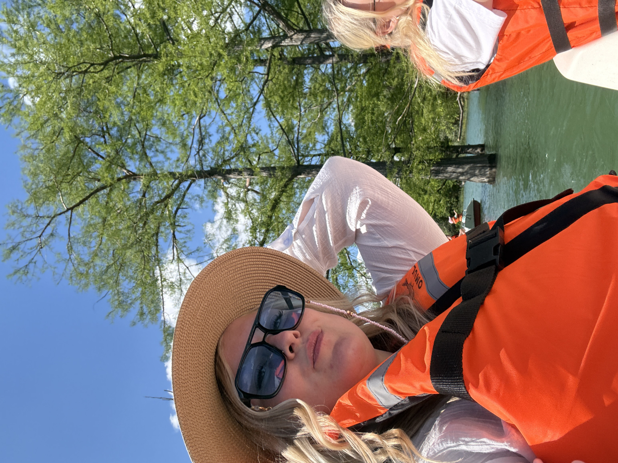 This photograph captures two individuals, likely women, enjoying a sunny day on a body of water in what appears to be Sukko, Russia, consistent with its famous Cypress Lake. The image is oriented vertically, presenting a dynamic perspective.
In the foreground, a blonde-haired woman is prominently featured, wearing a wide-brimmed straw hat and stylish black sunglasses. She has on a white long-sleeved top and a bright orange life vest, on which the text "OMWO" is visible. Her expression is relaxed and calm as she looks towards the camera. Her blonde hair falls in loose waves around her face and shoulders.
Partially visible in the upper right of the frame, another person with blonde hair can be seen, also wearing a white top and an orange life vest, suggesting they are on the same boat or nearby.
The background is dominated by a clear blue sky with fluffy white clouds, interspersed with lush green trees featuring slender branches. These trees appear to be growing directly out of the tranquil, greenish-blue water, a distinctive characteristic of the cypress trees found in Sukko Lake. The calm water surface reflects the surrounding greenery and sky. A white boat hull is partially visible in the upper right, suggesting they are aboard a vessel.
The overall impression is one of leisure and relaxation amidst a picturesque natural setting, likely during a boating excursion on a beautiful day.