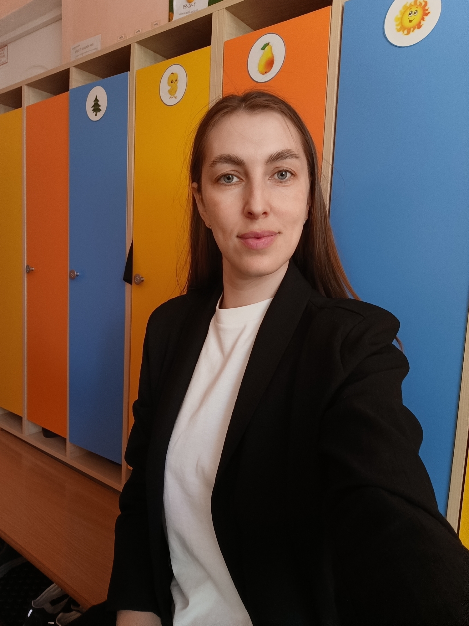 A woman is posing for a selfie indoors. She has long brown hair, fair skin, and is wearing a white t-shirt and a black blazer. Behind her are lockers with alternating yellow, orange, and blue doors. Each locker door has a circular sticker with an image on it: a yellow tree, a yellow chick, a yellow pear, and a yellow sun. The lockers are in a room in Chaikovsky, Russia. The image conveys a sense of everyday life and the setting is likely a school or kindergarten.