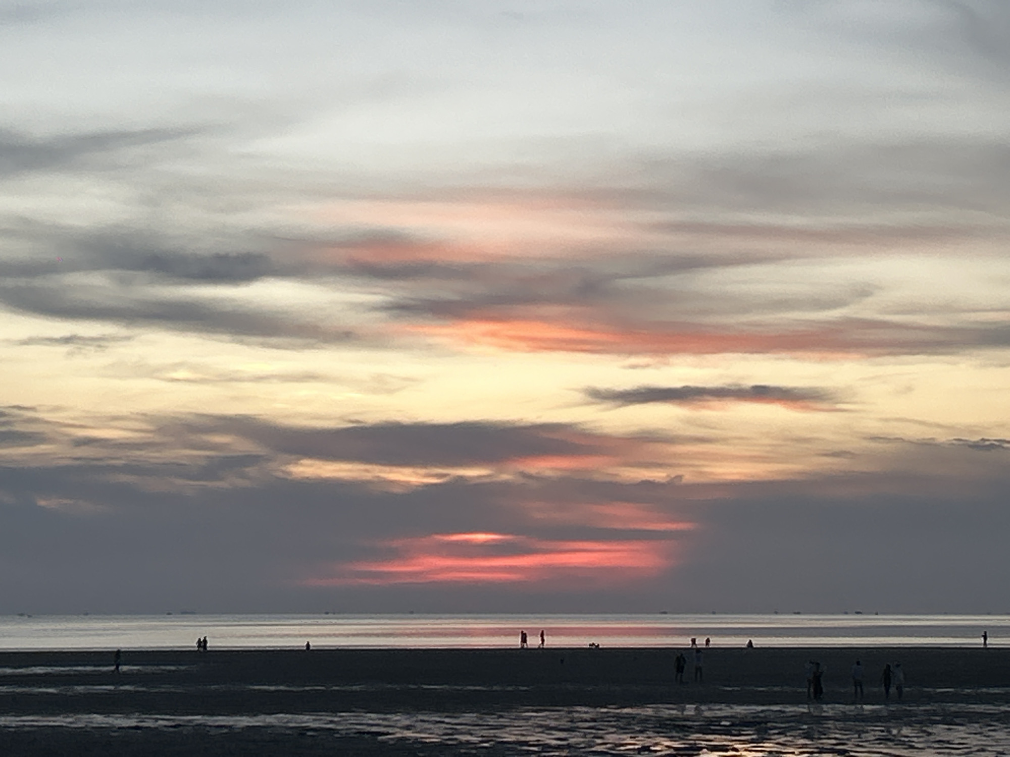 The image depicts a sunset over the ocean at Baan Sri Thanu, Thailand. The sky is filled with striated clouds, colored grey, yellow, and pink, closest to the horizon. The horizon is barely visible and appears to separate the sky from the calm ocean. In the foreground, wet sand reflects the sunset colors. Several figures are scattered across the beach, appearing as dark silhouettes against the illuminated sky and water.