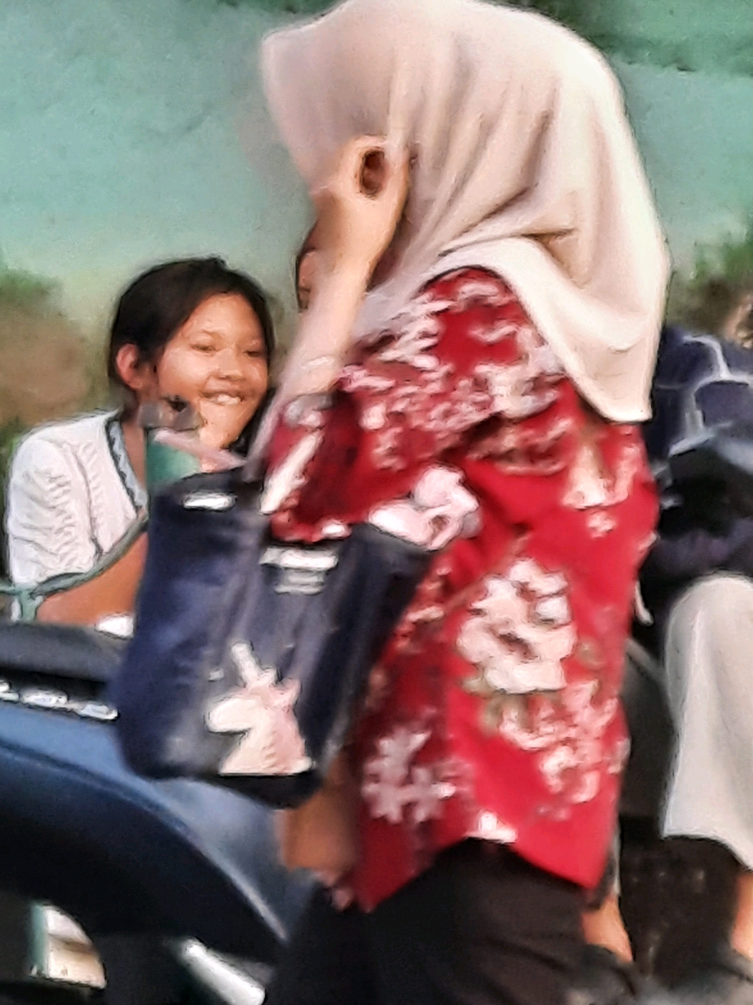 In East Jakarta, Indonesia, a young girl with dark hair and a white shirt smiles as she sits on a motorcycle. Next to her, a woman wearing a light-colored hijab and a red floral-patterned shirt is partially visible, her hand touching her face. A dark blue bag with a unicorn decal hangs from the motorcycle. The background is a blurred green and teal, suggesting an outdoor environment.