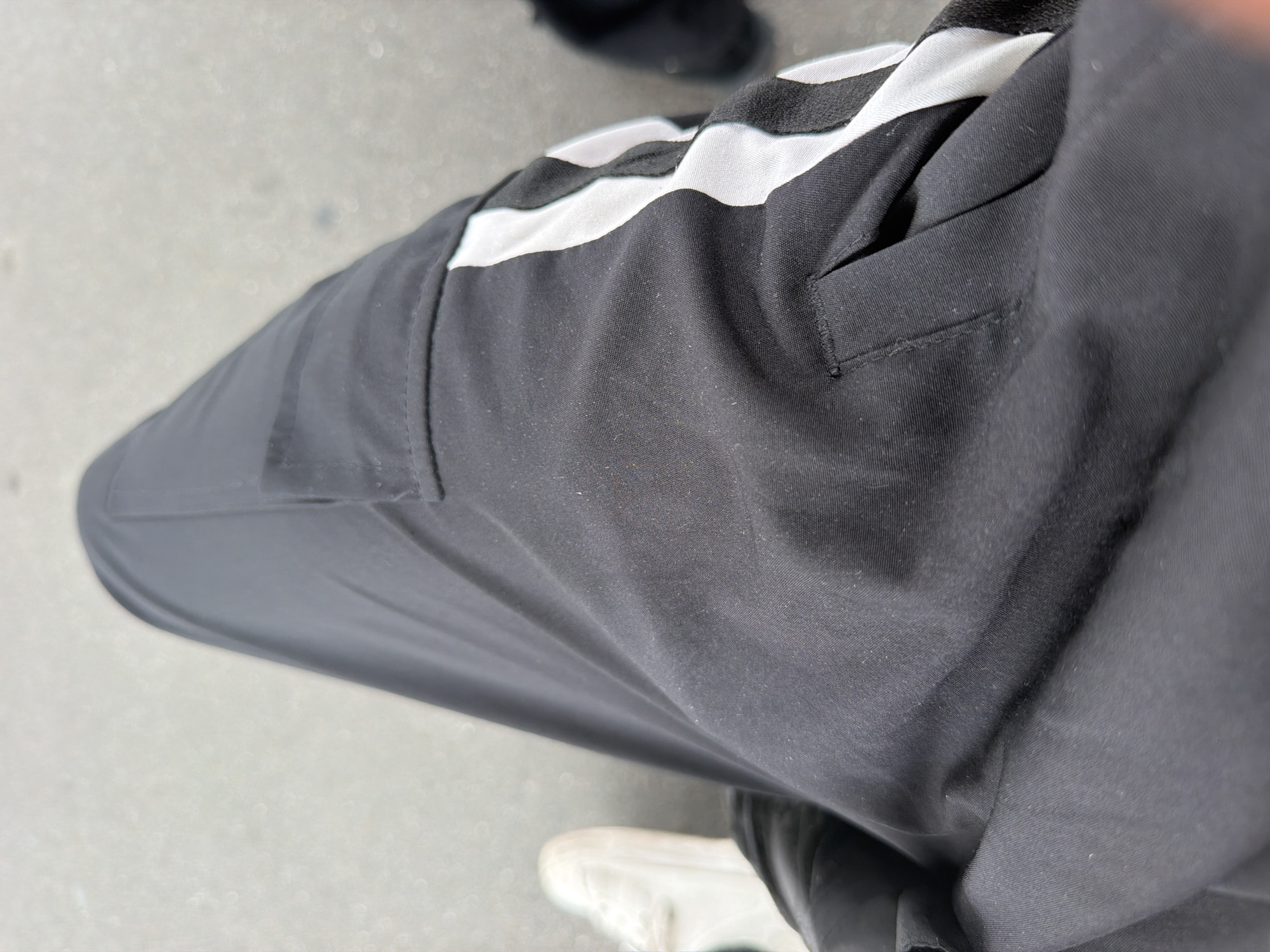 The image shows a close-up view of a person's legs wearing black athletic pants with white stripes on the side. A white sneaker is visible on the left side of the frame. The background is blurred and appears to be a paved surface, possibly a sidewalk or road. The scene is outdoors. The specific location within Adachi, Japan, cannot be determined from the visual information provided.