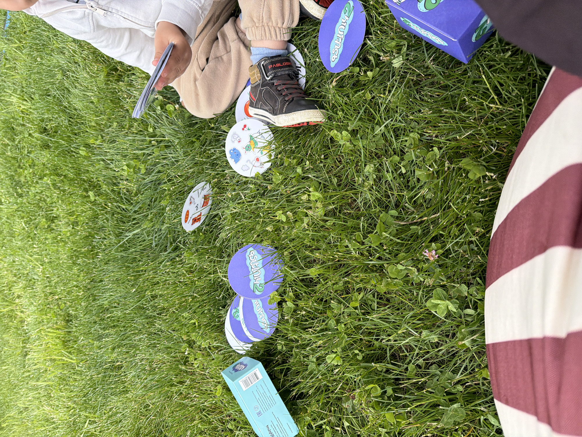 A child sits on a patch of green grass strewn with clover, likely in an outdoor space. The child is wearing tan pants, a white jacket, and black shoes with red laces. They hold a stack of cards in their hand. Several circular cards with cartoon images are scattered around, some lying under the child's shoe. A blue box and a purple box are also on the grass. The presence of a striped object in the frame is evident. The location is potentially in Chkalov, Armenia.