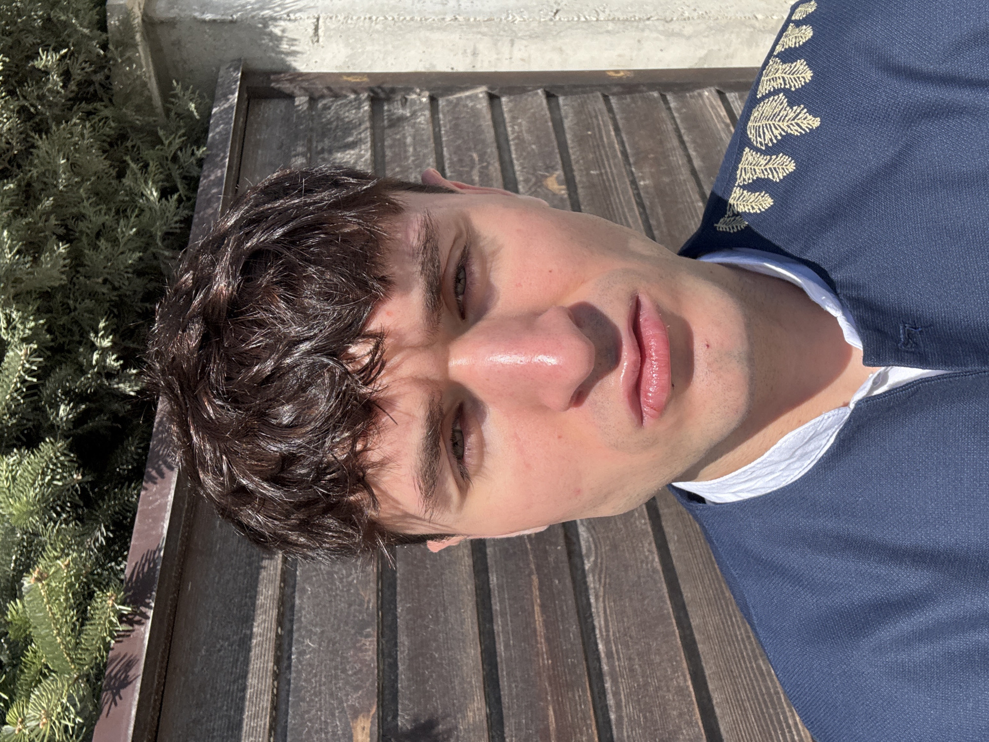 The image shows a close-up of a man lying on a wooden bench in Bania, Bulgaria. He is wearing a blue collared shirt with white trim and embroidered gold leaf detailing on the shoulder. The man has short, dark curly hair and fair skin. The lighting indicates it is likely daytime. Part of a green bush is visible in the left corner of the image. A concrete structure is also visible at the top of the frame.