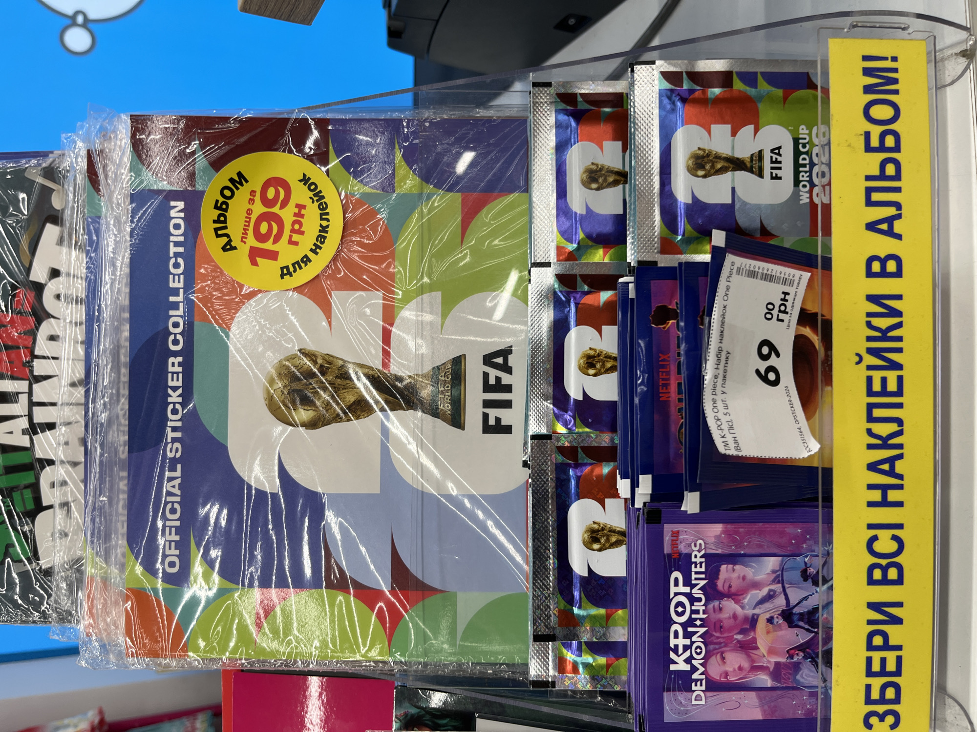 The image shows a close-up view of a retail display stand in a store, likely in Kharkiv, Ukraine, as indicated by the Ukrainian text on the signage. The display features several items related to sticker collections.
At the top, a large album titled "OFFICIAL STICKER COLLECTION" is prominently displayed. It features a graphic of the FIFA World Cup trophy and the text "FIFA WORLD CUP 2026". A yellow circular sticker on this album reads "Альбом лише за 199 грн для наклеек", which translates to "Album for only 199 UAH for stickers".
Below the album, rows of smaller sticker packs are visible. These include packs with images of characters labeled "KPOP DEMON HUNTERS" and "NETFLIX". Another set of sticker packs features the FIFA World Cup trophy and the year "2026".
A bright yellow sign at the bottom of the display has large blue Ukrainian text that reads "ЗБЕРИ ВСІ НАКЛЕЙКИ В АЛЬБОМ!" which translates to "Collect all stickers in the album!".
The environment appears to be a well-lit indoor retail space, with the display stand made of clear plastic. There are no people clearly visible in the frame. The items are neatly arranged for sale.