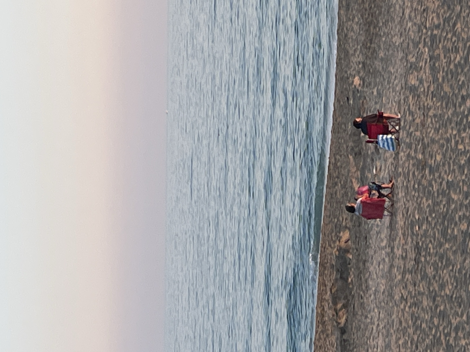 This media file captures a tranquil beach scene in Alanya, Turkey, presented in a sideways orientation. When viewed correctly as a landscape, the left side of the frame features the calm, blue-gray sea with subtle ripples across its surface. The right side is a sandy beach with dark, coarse sand. The sky, visible at the top, is light and hazy, with a faint pinkish-beige tint, suggesting the soft light of an overcast day or late afternoon/early evening.
Two individuals are relaxing on the beach. One person, closer to the foreground, is seated in a red portable beach chair, wearing a dark top. A blue and white striped object, possibly a bag or towel, rests beside their chair. Further back, another individual is also seated in a red beach chair, wearing a pink top and dark shorts, with their feet visible. The scene portrays a peaceful moment of leisure by the sea. There is no visible text in the image.
