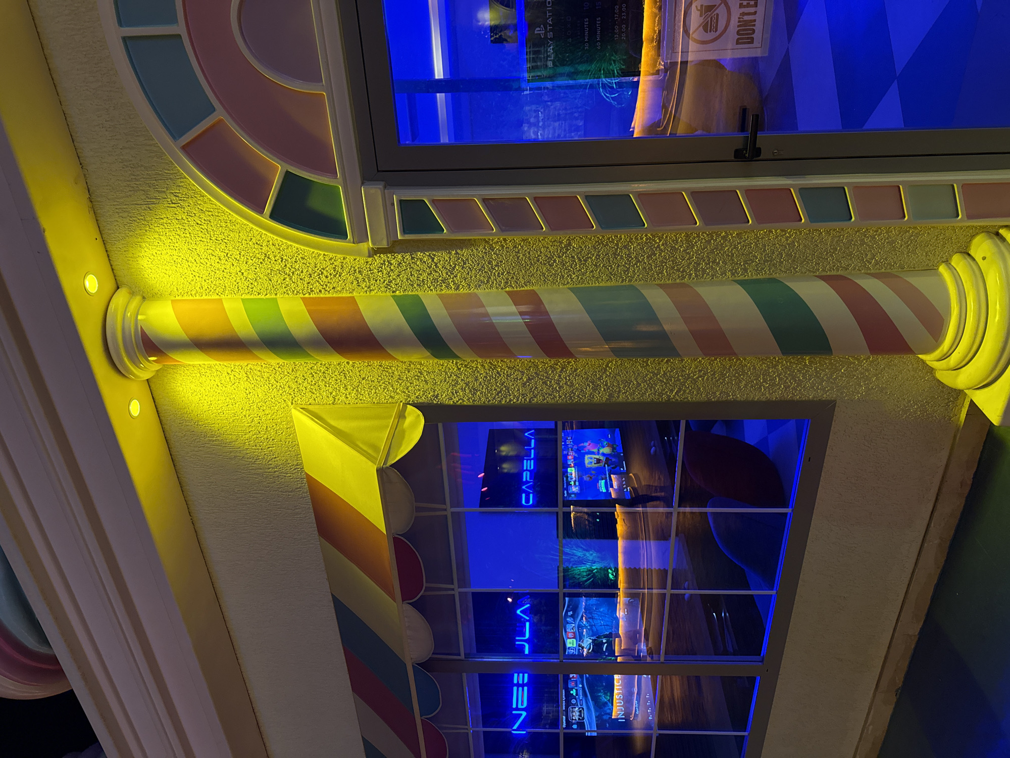 The image shows the exterior of a brightly lit establishment, likely a gaming or entertainment venue, in Bodrum, Turkey. The facade features a vibrant candy-striped column and colorful decorative accents around a glass door and windows. Neon signs are visible through the windows, with one appearing to read "INEJ ULA" and another "CAPELLA." Inside, multiple screens display video games, including one labeled "INJUSTICE." The blue interior lighting casts a strong glow onto the exterior. A "DON'T EAT" sign with a crossed-out burger is affixed to the glass door. The overall impression is one of a playful and energetic commercial space.