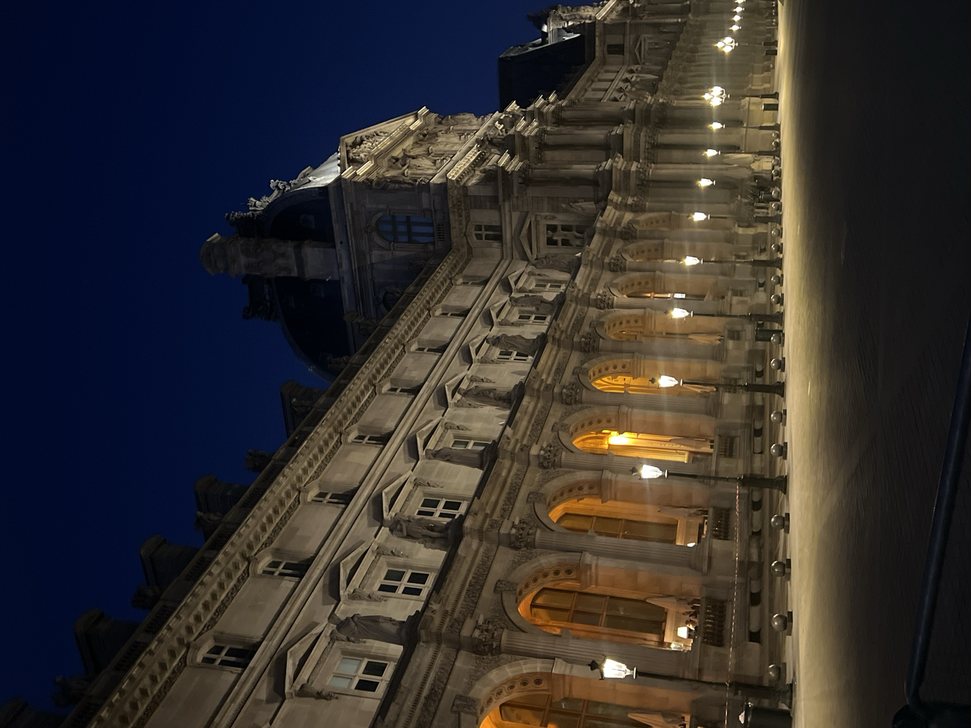 The image shows the Louvre Museum in Paris, France at night. The grand building, illuminated by streetlights, is captured from a low angle, emphasizing its imposing architecture. The sky above is a deep, dark blue, indicative of nighttime. The foreground features a paved area with evenly spaced spherical bollards and a line of classic Parisian streetlamps casting warm light onto the ground. The windows of the museum are glowing with a warm, interior light, and some canopies are visible in the arcaded sections. No people are clearly visible in the frame. The scene conveys a sense of quiet grandeur and historical presence.