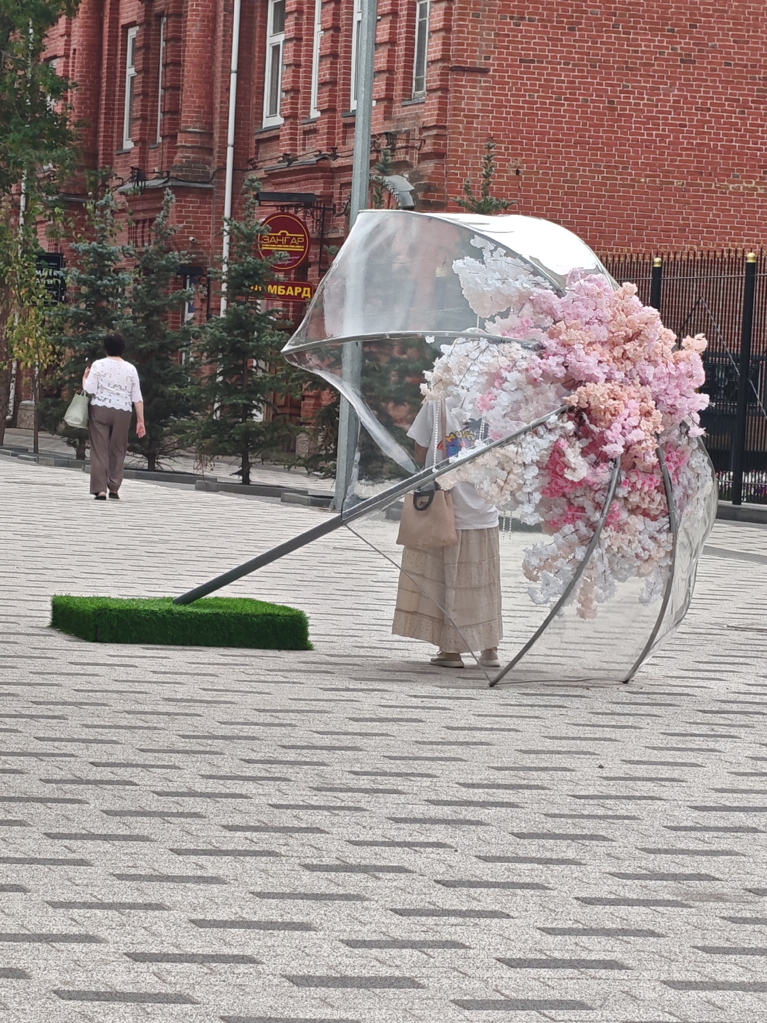 In Petropavl, Kazakhstan, a street scene unfolds with a brick building serving as a backdrop. A woman, dressed in a white top and brown trousers, walks away from the viewer on a patterned paved surface. In the foreground, a large, clear umbrella lies on its side. The umbrella is adorned with a dense arrangement of artificial flowers in shades of pink, white, and light orange, creating a vibrant floral display. A small, rectangular patch of artificial green grass is positioned near the base of the umbrella's handle, suggesting an artistic installation or a decorative element. The overall atmosphere is that of an outdoor urban setting, possibly a public square or promenade.