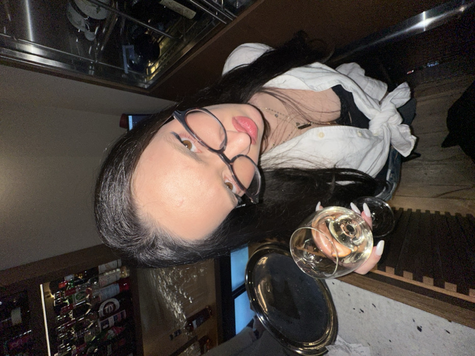 An overhead, wide-angle image depicts a young adult female within an establishment reported to be in Yerevan, Armenia. The subject, possessing long dark hair, wears dark-framed glasses, a white button-down shirt tied at the waist over a dark undergarment, and multiple necklaces. She holds a stemmed glass containing a clear, light-colored liquid. Her facial expression includes slightly pursed lips and a direct gaze towards the camera. To her left, a bar counter is visible with a silver oval-shaped tray and, in the background, shelves stocked with numerous liquor bottles. To her right, a tall wooden cabinet with a glass front displays multiple wine bottles, suggesting a wine cooler or display. The overall lighting is dim, indicative of an evening setting in a bar or restaurant.