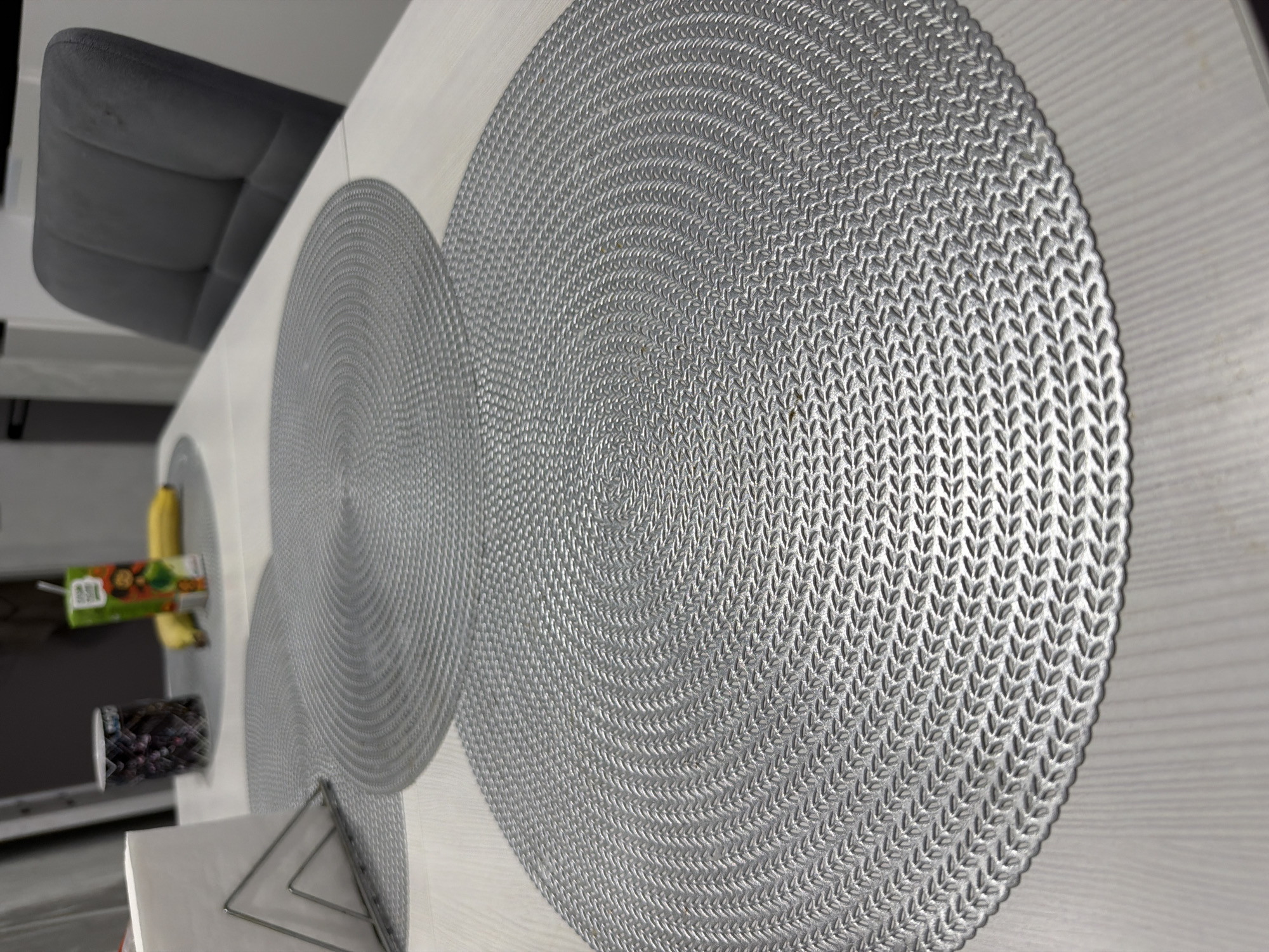 The image shows a close-up of a table in what appears to be an indoor setting. The table has a light-colored surface with several silver, circular placemats on it. The placemats have a woven texture. There is a chair with a gray upholstered back visible along one side of the table. On the opposite side of the table, items such as a juice box, a jar of fruit or candy, and a triangular object are visible, suggesting this may be a kitchen or dining area. There's nothing to definitively link the location to Chaikovsky, Russia.