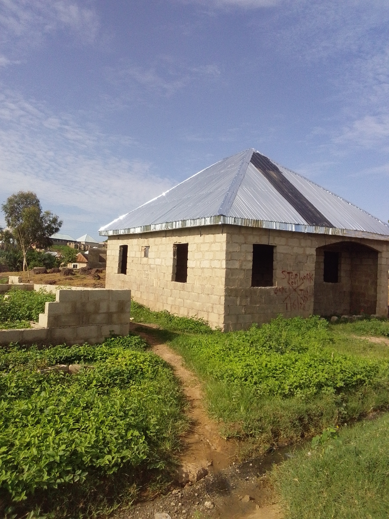 The image depicts an unfinished single-story residential structure made of light-colored concrete blocks, situated in a developing area within Keffi, Nigeria, under a bright, partly cloudy sky. The building features a newly installed multi-pitched roof composed of corrugated metallic sheets, with a prominent dark-colored strip along the central ridge. Rectangular openings for windows and an arched entry point are present, but no doors or window frames are installed. Red graffiti is visible on the front wall, including the phrases "STOP WORK" and other illegible markings. The foreground consists of a dirt path winding through dense green vegetation, with a small drainage ditch or eroded channel to the right. To the left of the central building, partial foundation walls of another unfinished structure are visible, surrounded by similar green foliage. In the midground, a large tree with sparse leaves stands on slightly elevated ground, and further in the background, other residential buildings and construction appear, indicating an expanding settlement. No individuals are present in the scene.