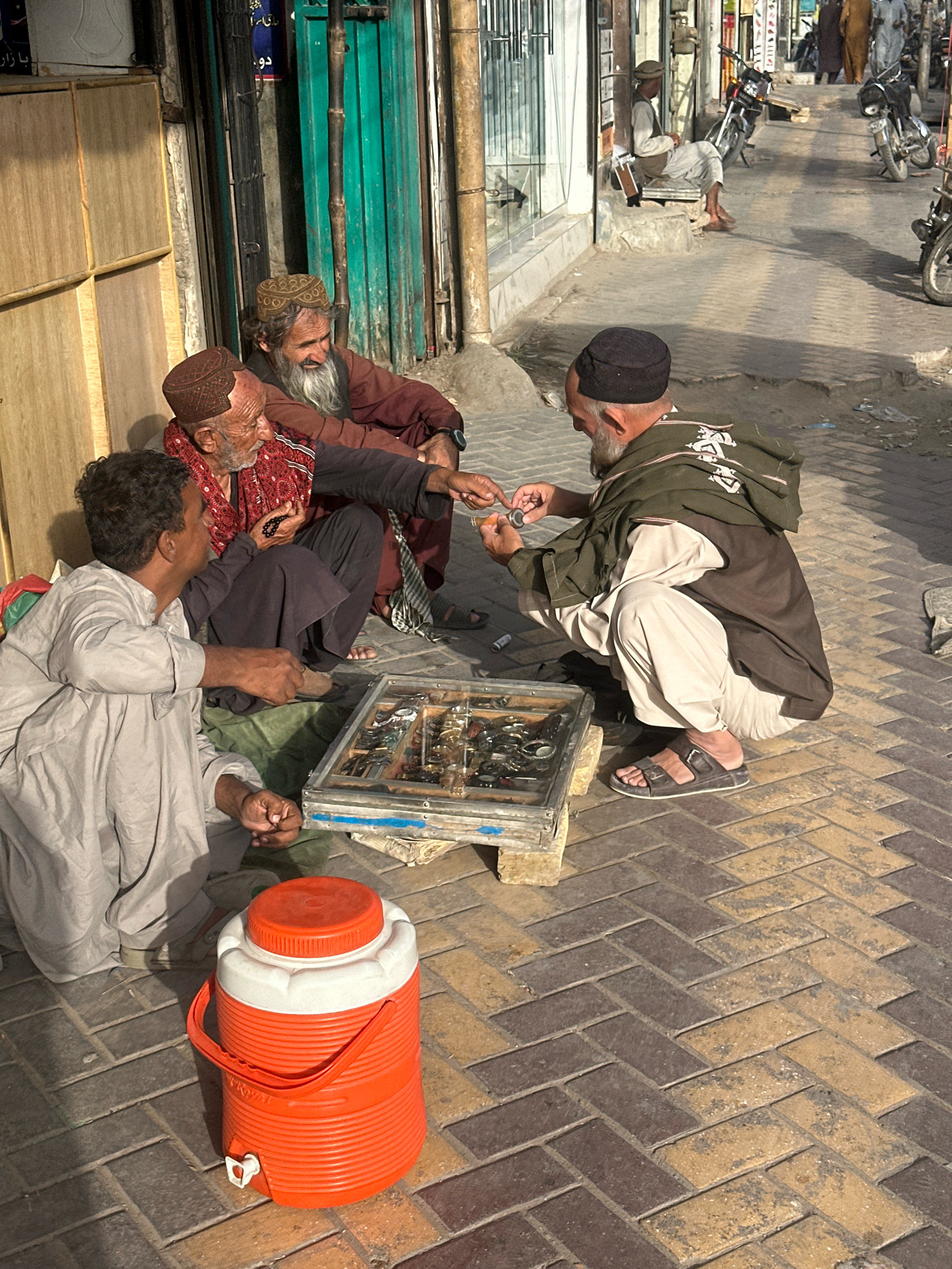 The image depicts an outdoor street scene in Quetta City Tehsil, Pakistan. Four men are engaged in social interaction around a display case of what appear to be watches or jewelry. One man, wearing a light gray shalwar kameez, is seated and looking towards the display. Another man, in a dark shalwar kameez and a brown shawl, is squatting and examining an item from the case, which is a metal-framed box with multiple compartments. A third man, in a maroon shalwar kameez and a patterned headscarf, is seated and holding a string of dark beads. A fourth man, wearing a black cap and a dark green shawl over a light-colored shalwar kameez, is also squatting and appears to be in conversation with the man examining the items from the case. In the foreground, an orange and white water cooler with a spigot is prominently placed on the brick-paved ground. The background shows a building with a green metal door and other structures, suggesting a commercial or market area. Motorcycles are visible in the background, indicating typical street activity. The lighting suggests it is daytime, possibly late afternoon.
