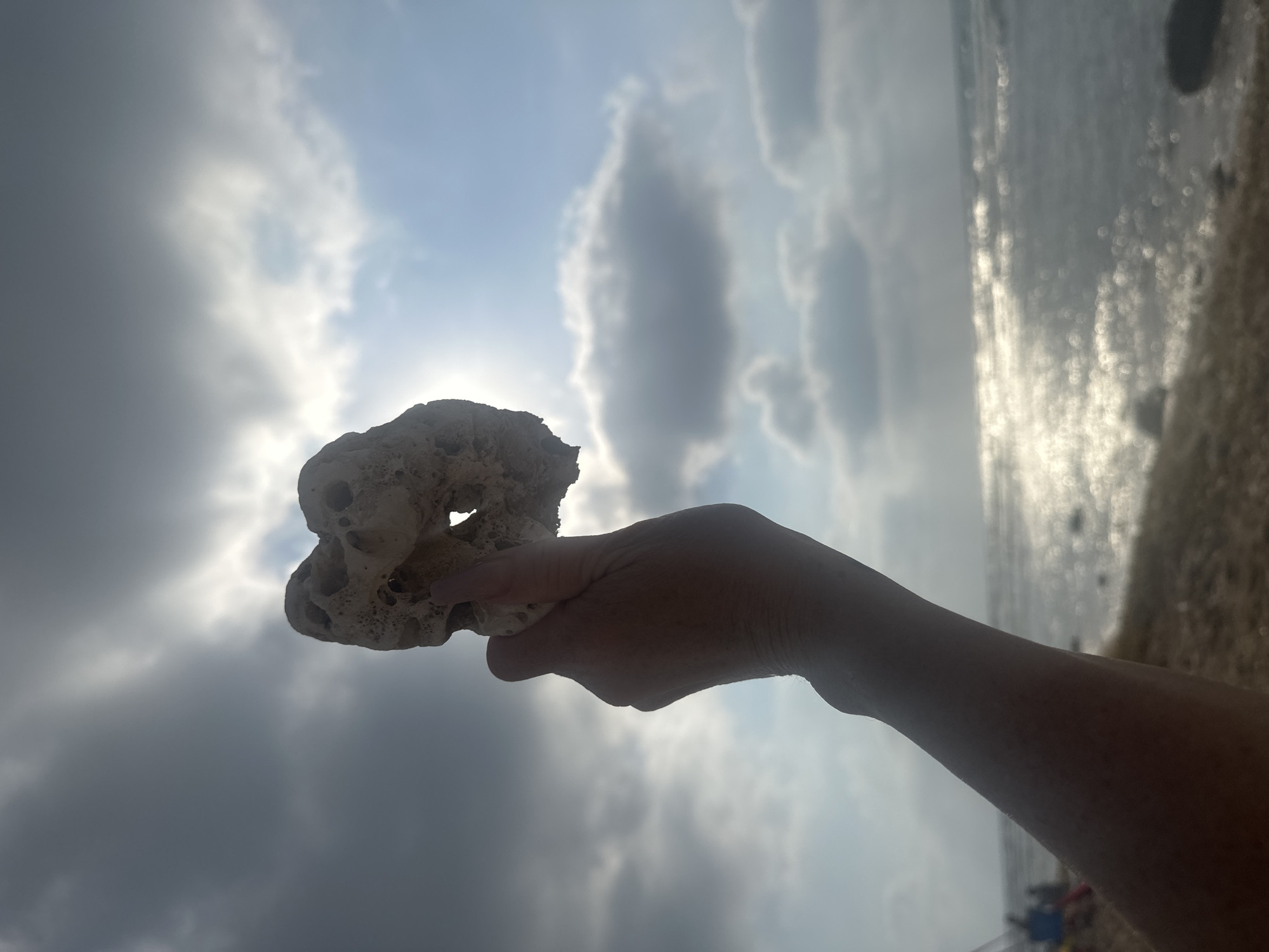 The image shows a person's hand holding a porous, light-colored rock against a sky with dark and light clouds. The sun is partially obscured by a cloud, creating a bright halo effect. The background depicts the sea with shimmering sunlight reflecting on the water. The scene appears to be a beach, as indicated by the sandy shore and the presence of blurred figures and beach equipment in the distance. The location is not specifically identifiable as being in Haifa, Israel, but the overall environment suggests a coastal area.