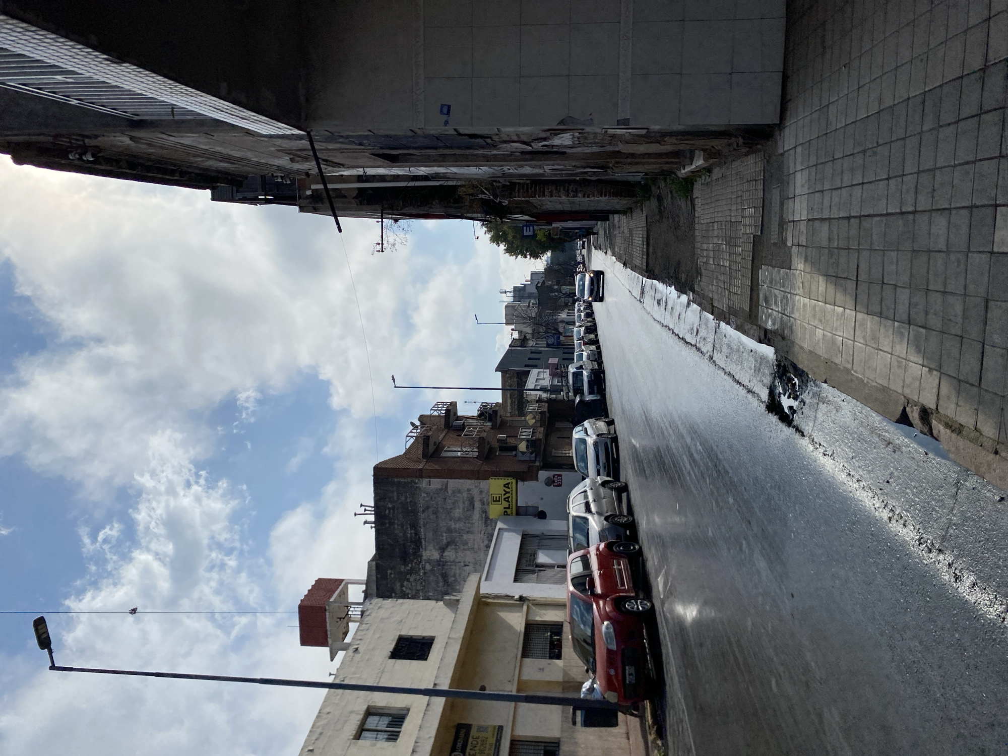 This image depicts a street scene in Cordoba, Argentina, likely on a cloudy day with the sun partially obscured. The perspective is from a sidewalk facing a wet asphalt road lined with parked cars. On the left side of the street, a white building with windows and a red-tiled water tank on its roof is visible, along with other older multi-story buildings. A yellow sign reading "E PLAYA ESTACIONAMIENTO" is attached to one of the buildings. Several cars, including a red hatchback, are parked along the curb. The road appears to have puddles, suggesting recent rain. On the right side of the image, the facade of a dilapidated building with peeling paint and cracked plaster dominates the foreground. A tiled sidewalk with some sections in disrepair runs alongside this building. Further down the street, more buildings and vehicles are visible, with a cluster of taller modern buildings in the distance. A street lamp with a light fixture is prominently positioned on the left side of the frame, extending upwards against the sky, where a wire with what appears to be a person or object suspended from it is visible.