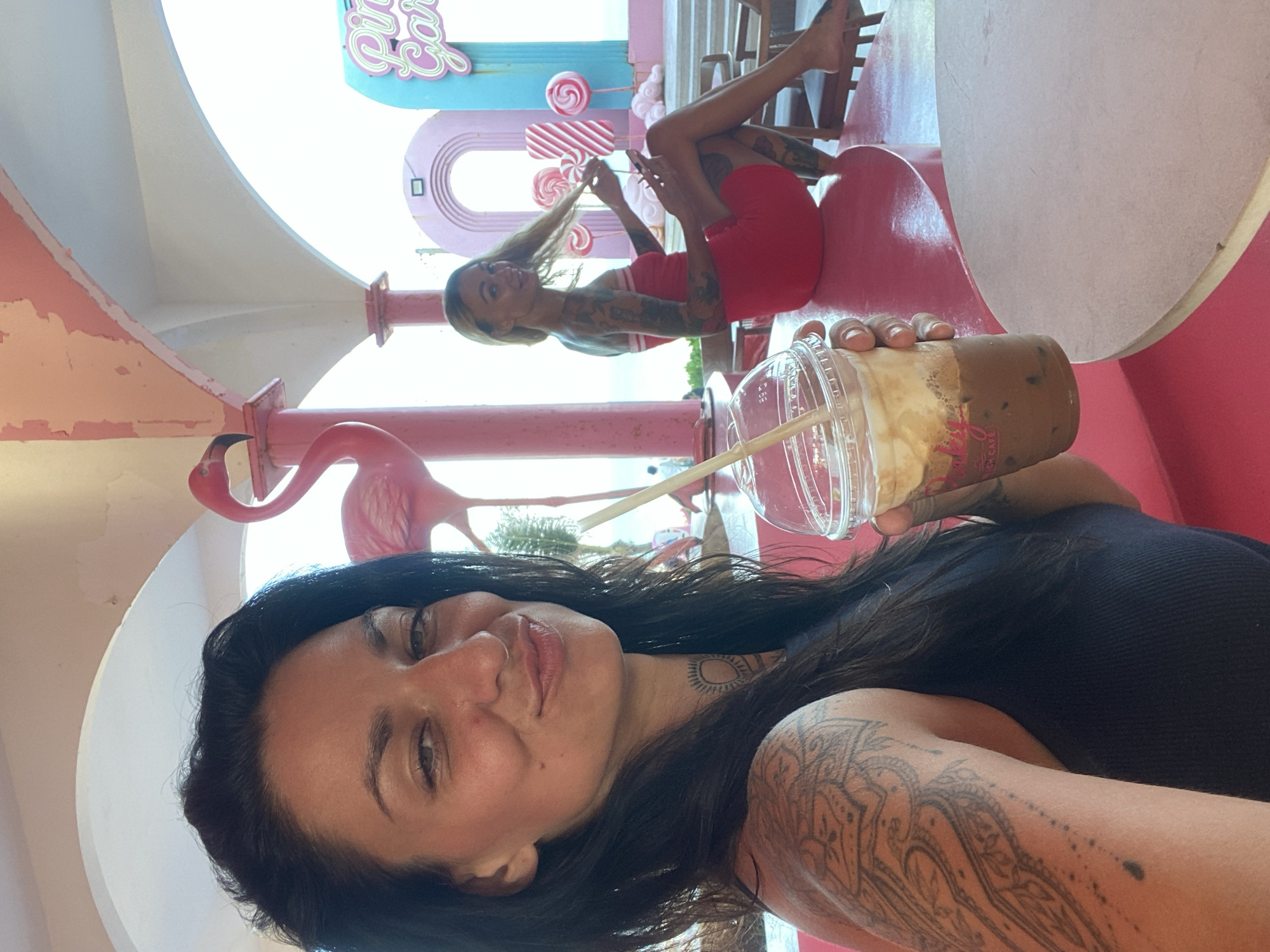 The image captures a selfie of a woman with dark hair and multiple tattoos, holding a beverage with a straw. She is smiling and looking towards the camera. In the background, another woman with blonde hair and tattoos is seated, wearing a red dress and playing with her hair. The environment is vibrantly decorated in pink and features a large pink flamingo statue, large candy-like decorations, and a neon sign that reads "Pink Garden." The setting appears to be an outdoor cafe or a themed attraction, possibly located in Tien Thanh Ward, Vietnam, given the context provided. The overall atmosphere is cheerful and colorful.
