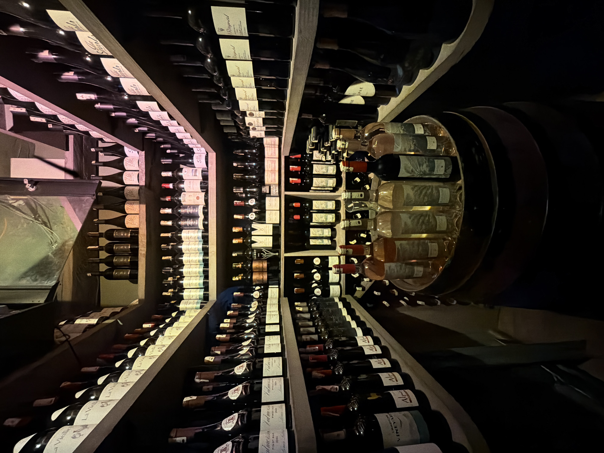 The image shows a dimly lit wine cellar with shelves filled with numerous bottles of wine. The bottles are predominantly dark glass with white and colored labels. The shelves are made of wood and are stacked in multiple tiers. In the foreground, a barrel is visible with several bottles of wine arranged on top, some of which appear to be rosé. The lighting is dramatic, casting deep shadows. The scene is devoid of people. The location is identified as being in the city of Marrakesh, Morocco, though there are no specific visual cues in the image to confirm this location.