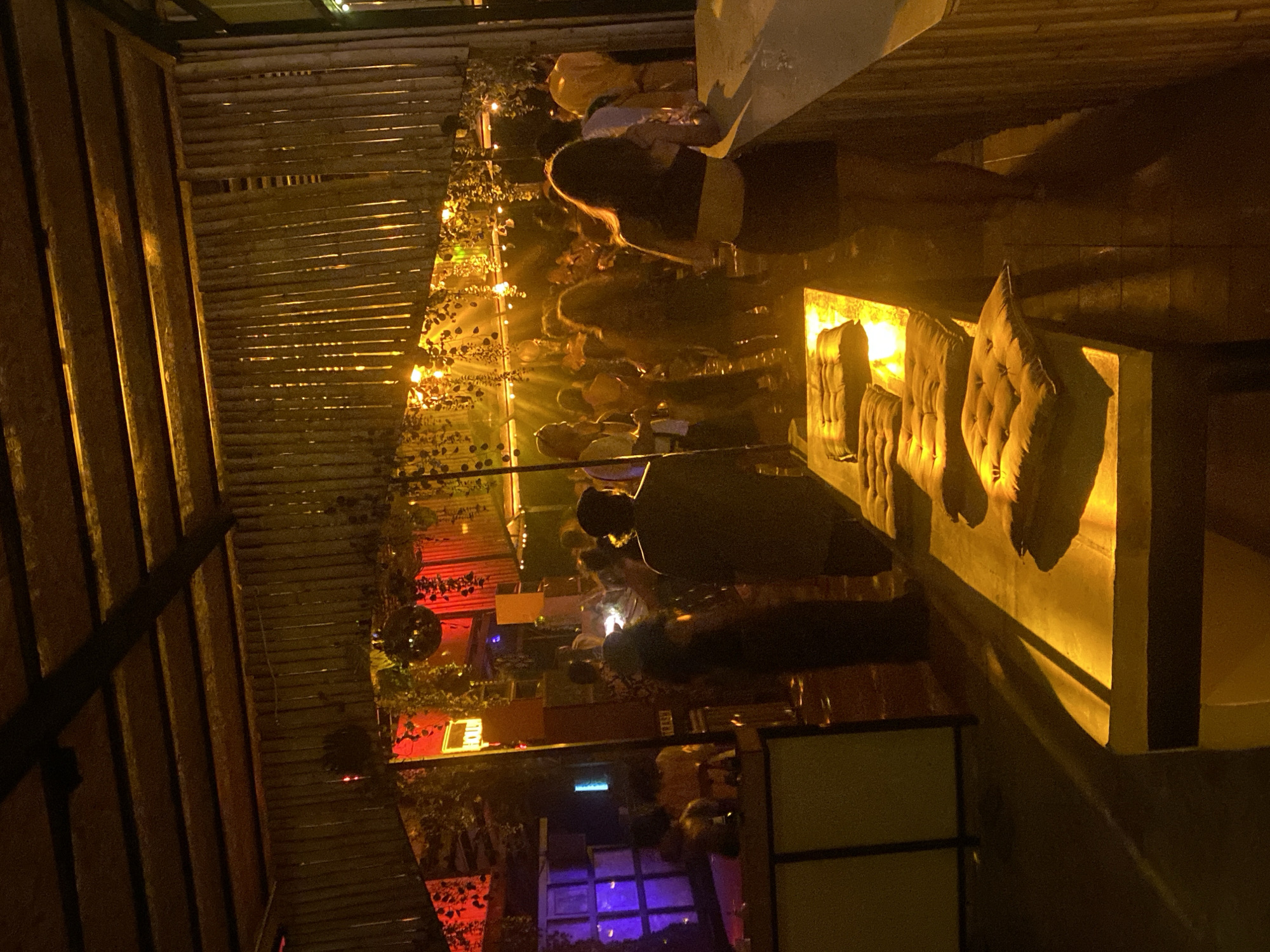 The scene appears to be a dimly lit outdoor restaurant or bar in Baan Hin Kong, Thailand, at night. The interior architecture features a wooden slat ceiling and walls on the left. Seating is visible on the right, with several tufted cushions lit by a warm, yellow light. Several people are mingling in the background, lit by string lights and colored lighting of red and blue. A disco ball hangs from the ceiling, adding to the festive atmosphere.