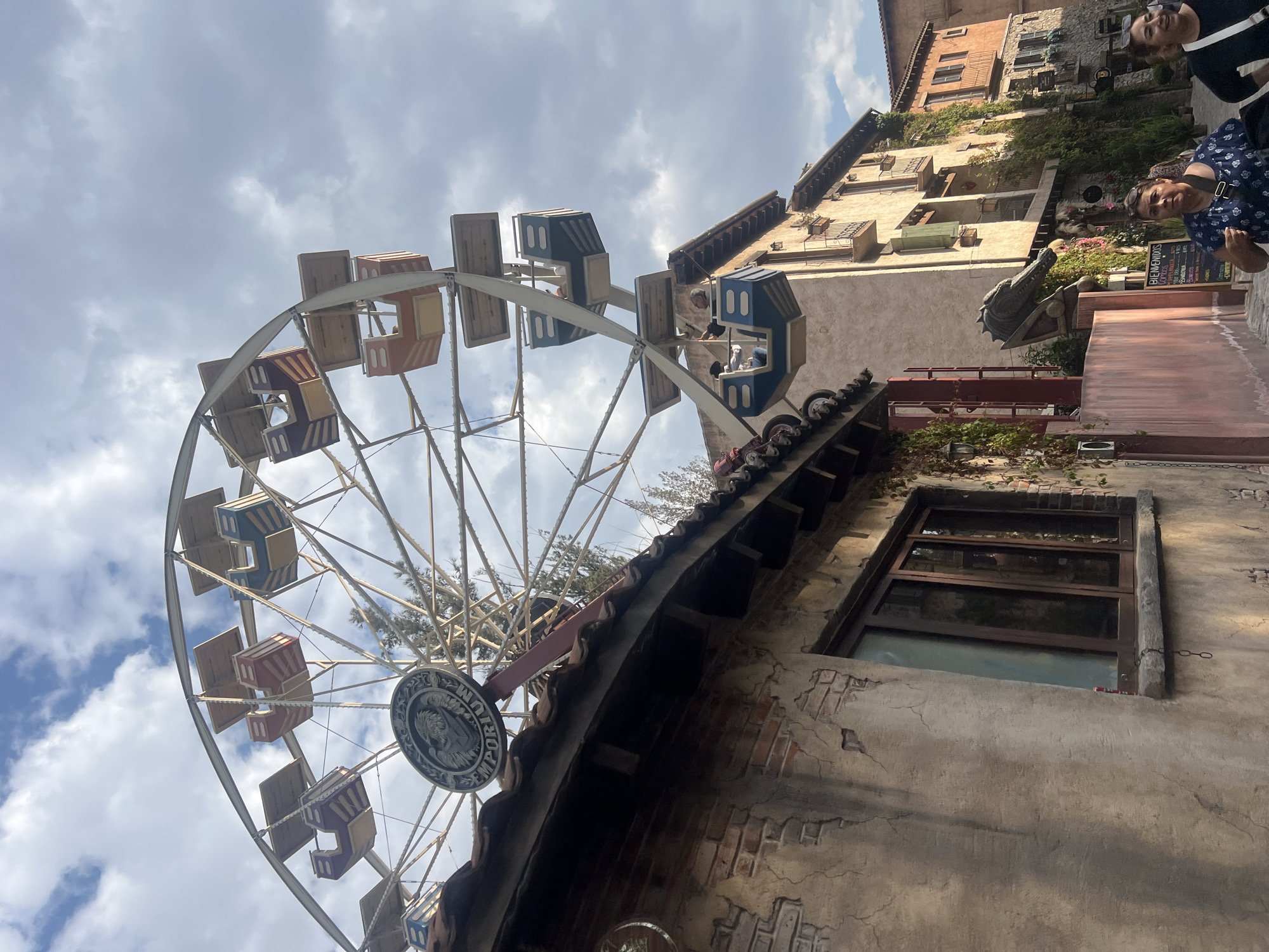 The image depicts a low-angle view of a Ferris wheel with colored cabins against a cloudy sky. The Ferris wheel is in Domingo Arenas, Mexico, situated beside aged buildings with tiled roofs and windows. Two individuals are visible in the upper right corner, one wearing a blue patterned shirt and the other partially obscured. A dragon statue is situated nearby. A menu board for a restaurant is positioned behind the statue. The buildings exhibit cracked walls and signs of aging.