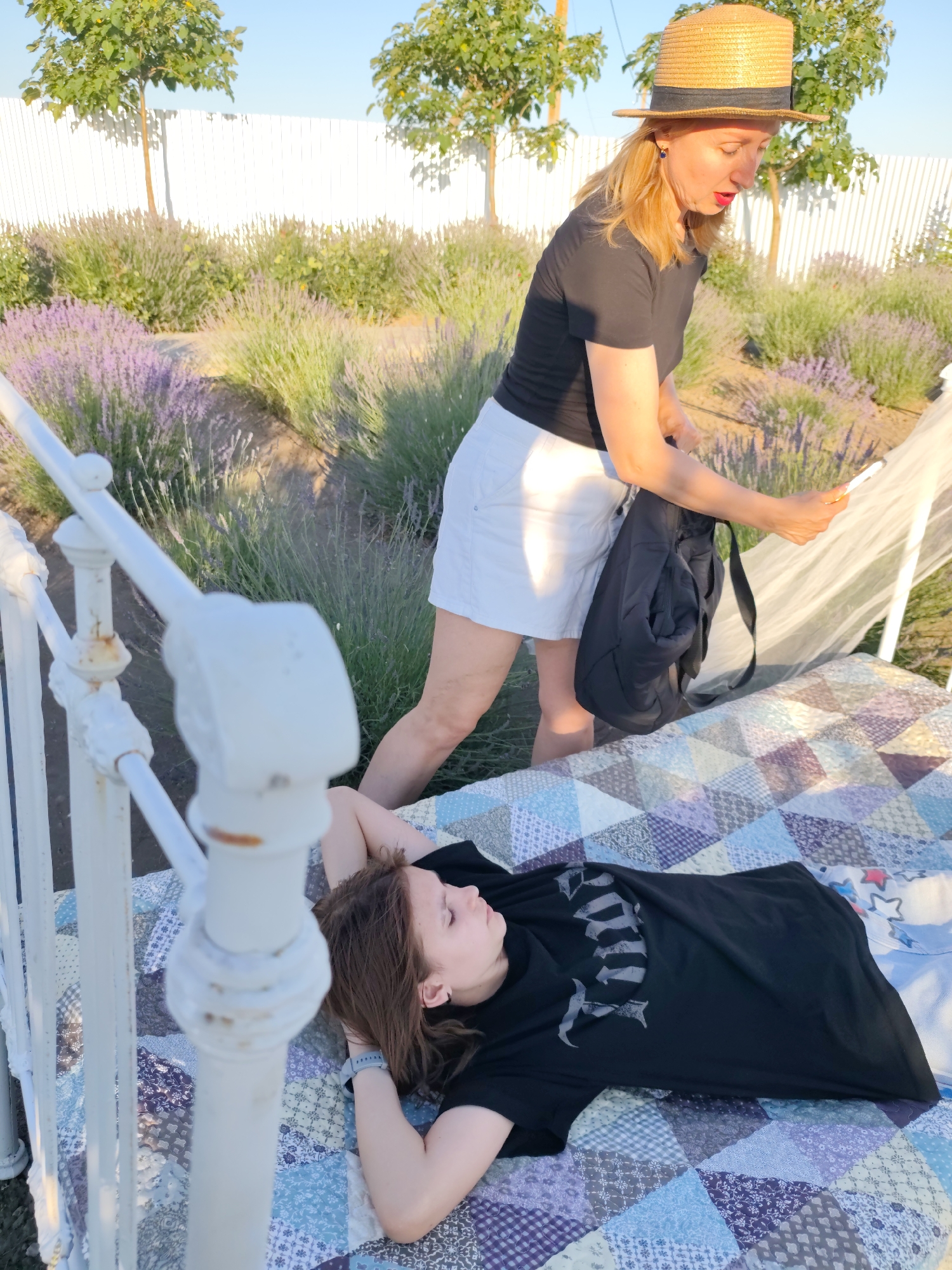 A woman wearing a straw hat and a black shirt stands over a young person lying on a quilt-covered bed outdoors. The woman is holding a phone and a black backpack. The bed appears to be part of an outdoor setup, possibly for lounging. Behind them, a field of lavender and trees are visible, with a white fence in the background. The scene is bathed in warm sunlight, suggesting it is late afternoon or early evening. The setting evokes a peaceful, rural atmosphere in Nechaiane, Ukraine. The young person is wearing a black t-shirt and a blue watch. The overall mood is relaxed and serene. There is no visible text in the image.