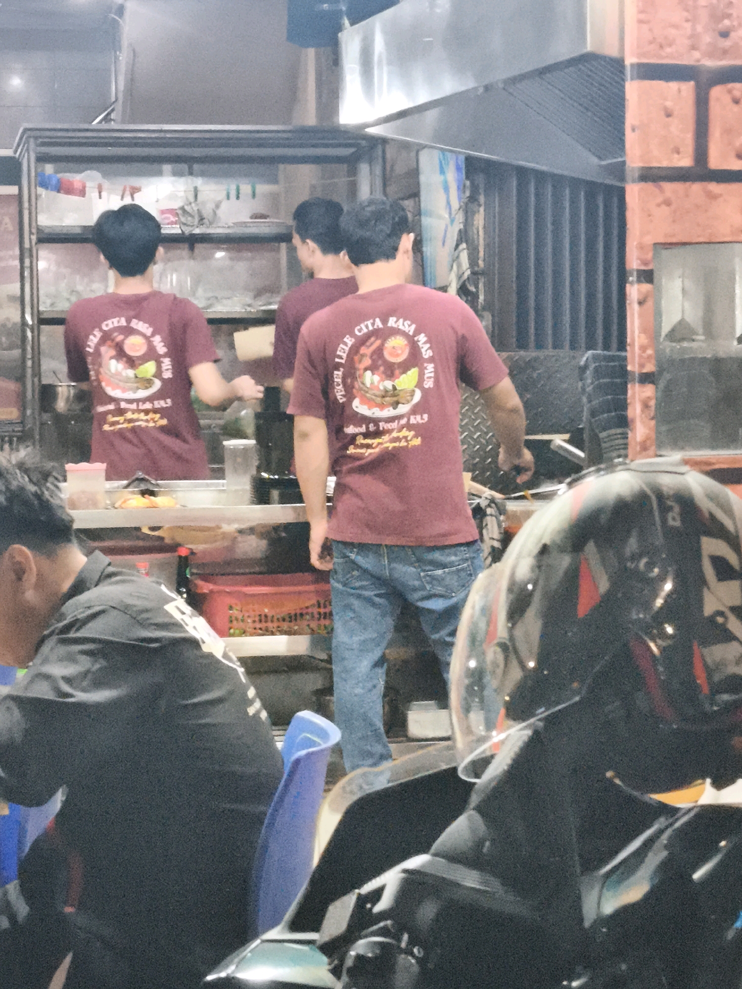 This is a medium shot, taken at a slightly low angle, of the exterior of a food stall in Palembang, Indonesia. The stall appears to be a popular spot, with several people working behind the counter and patrons seated at tables. In the foreground, a person wearing a black shirt is seated on a motorcycle, looking away from the camera. The person's face is partially obscured by a helmet. Behind the person, a young man wearing a maroon t-shirt with a graphic print is standing at the counter, looking down at the food being prepared. The t-shirt has text that appears to be in Indonesian. Another young man, also wearing a maroon t-shirt, is visible in the background, also working at the counter. The stall itself is brightly lit and features a variety of food items on display. The overall impression is of a busy and lively food establishment in the heart of Palembang.