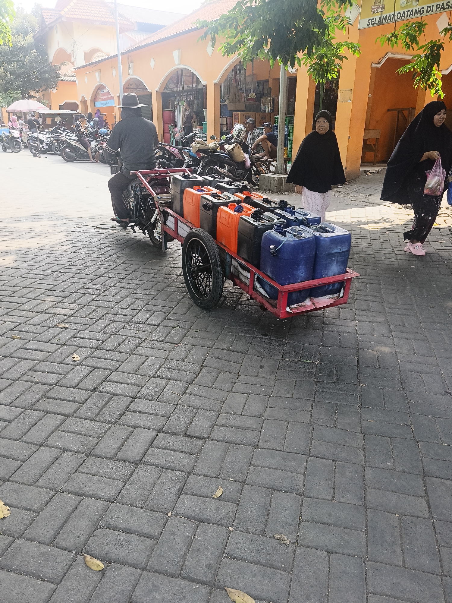 In a marketplace in Wedi, Indonesia, a man wearing a wide-brimmed hat and dark clothing rides a motorcycle that is towing a cart filled with multiple large, colorful plastic containers. The cart holds several orange and blue jerrycans, some of which appear to be full. Behind the motorcycle, two women are walking. The woman on the left, wearing a dark headscarf and black outer garment, walks with her head down. The woman on the right, also in a black headscarf and garment, carries a plastic bag and smiles as she walks. The ground is paved with grey interlocking bricks. In the background, there are stalls with awnings and trees. A sign above one of the stalls reads "SELAMAT DATANG DI PAS".