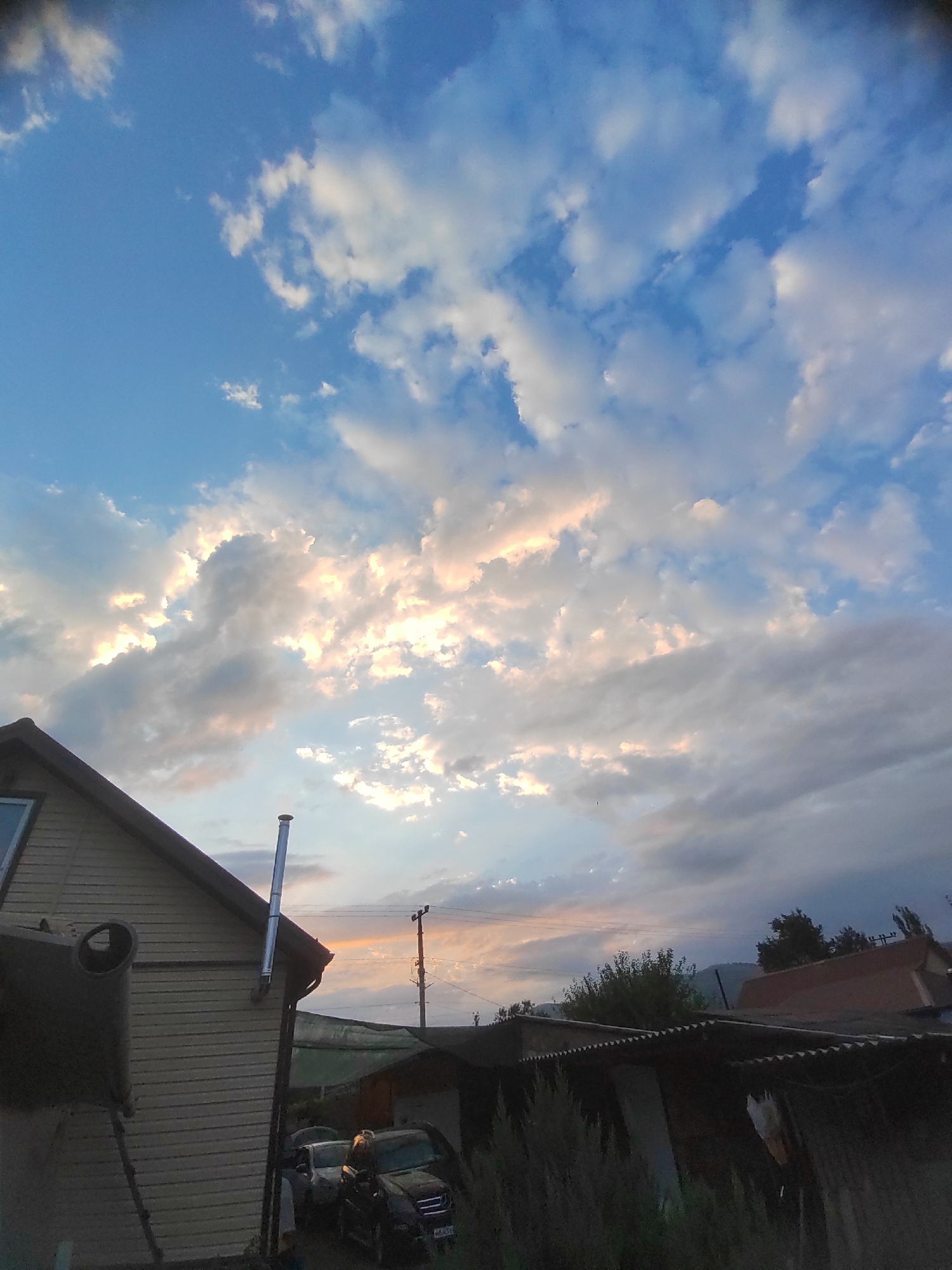 This is a wide-angle, eye-level shot taken outdoors during the day, likely in the city of Psebai, Russia. The scene is dominated by a sky filled with scattered clouds, some of which are illuminated by sunlight, creating a warm glow. In the foreground, a portion of a light-colored, horizontally paneled house with a dark roof is visible on the left. A metal chimney pipe extends vertically from the roof. In the middle ground, to the right of the house, there are several dark-colored structures with corrugated roofs, one of which appears to be a garage or shed. Two cars are parked in front of these structures. A power line pole with wires is visible in the background. The overall environment suggests a residential or semi-rural setting.