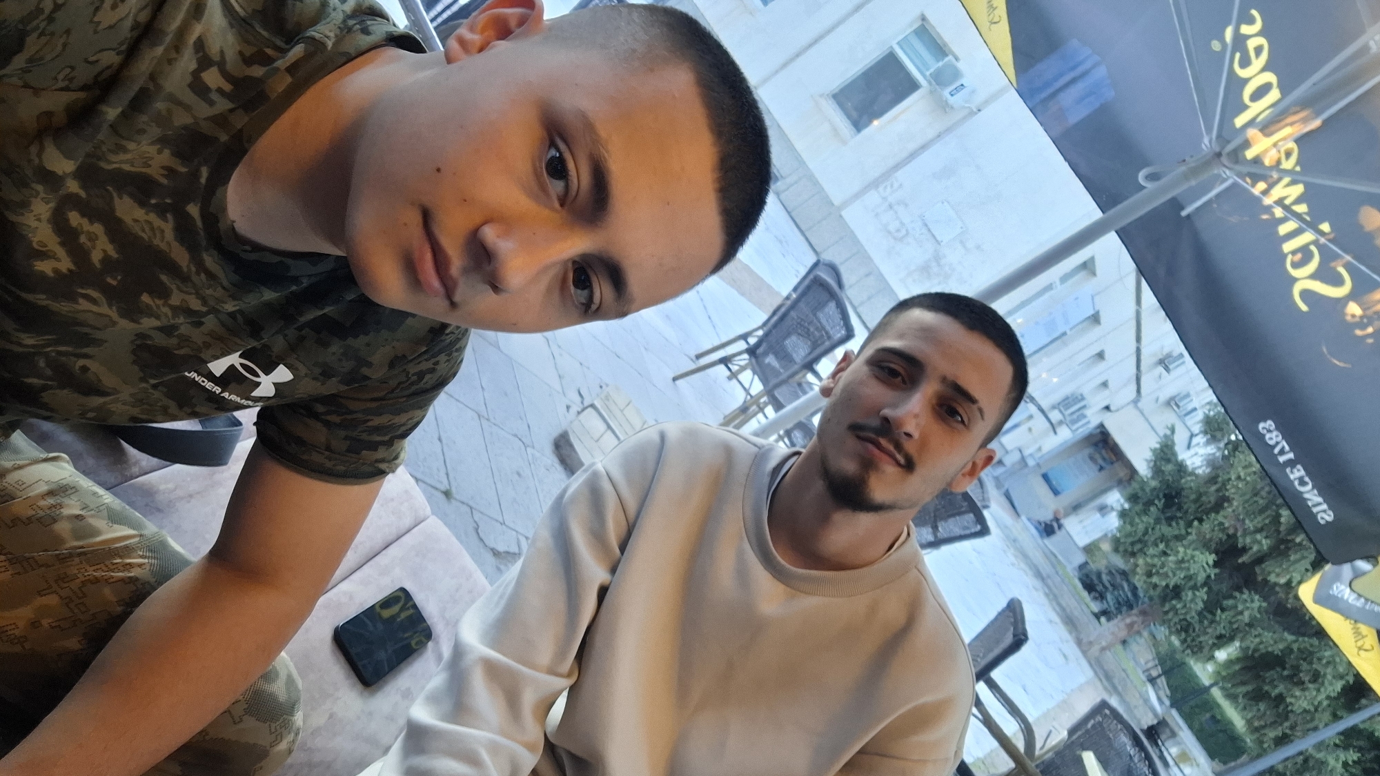 Two young men are seated at an outdoor cafe. One wears a camouflage t-shirt, and the other wears a tan sweatshirt. A smartphone is on the table near the man in camouflage. The outdoor cafe is in Svishtov, Bulgaria.