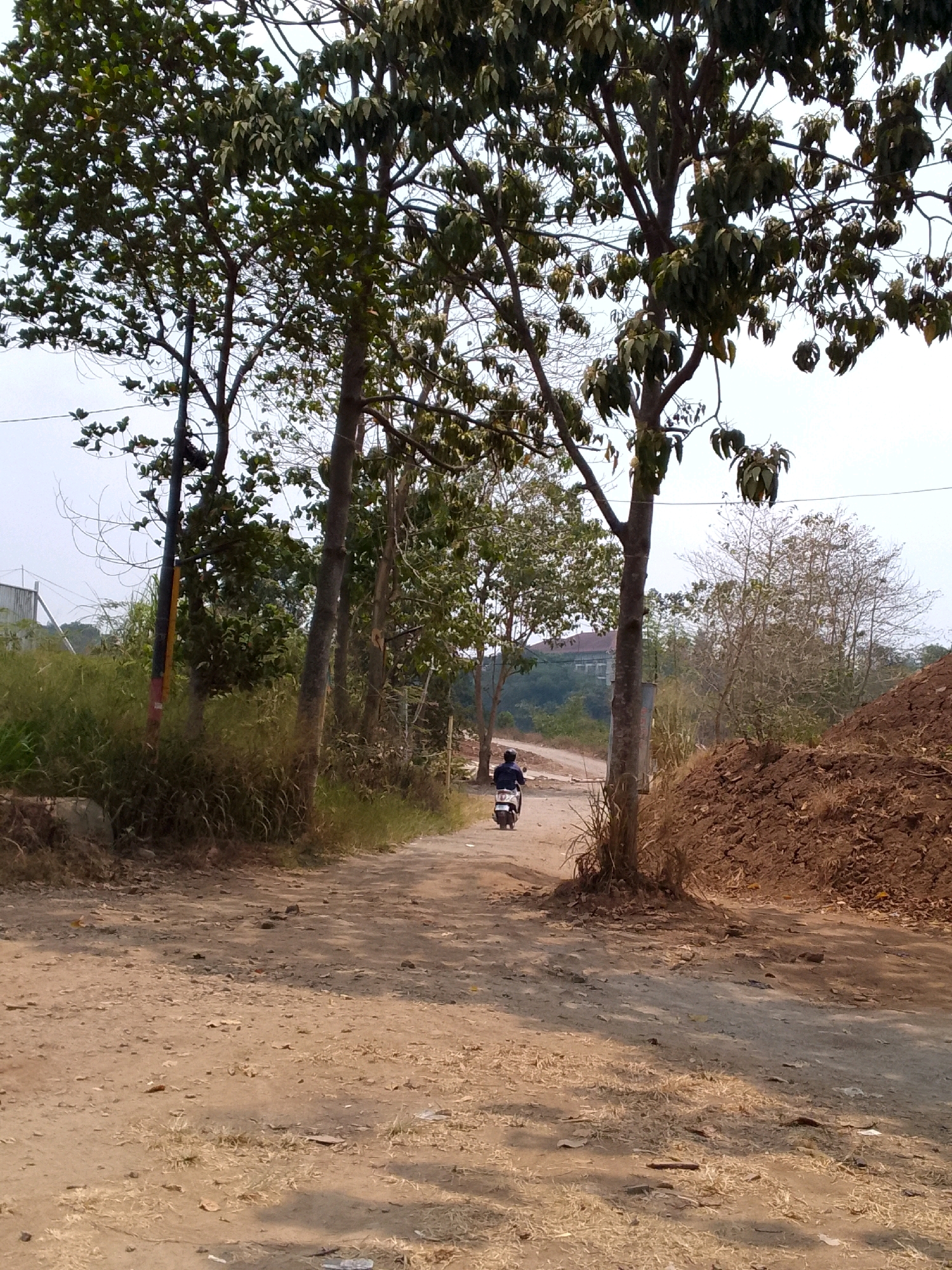 A dirt road winds through a semi-rural area, lined with trees and vegetation. In the distance, a person on a scooter is riding away from the camera down the road. To the right of the road, a large pile of soil or dirt has been accumulated. The sky is overcast. The scene appears to be in Cipeundeuy, Indonesia.
