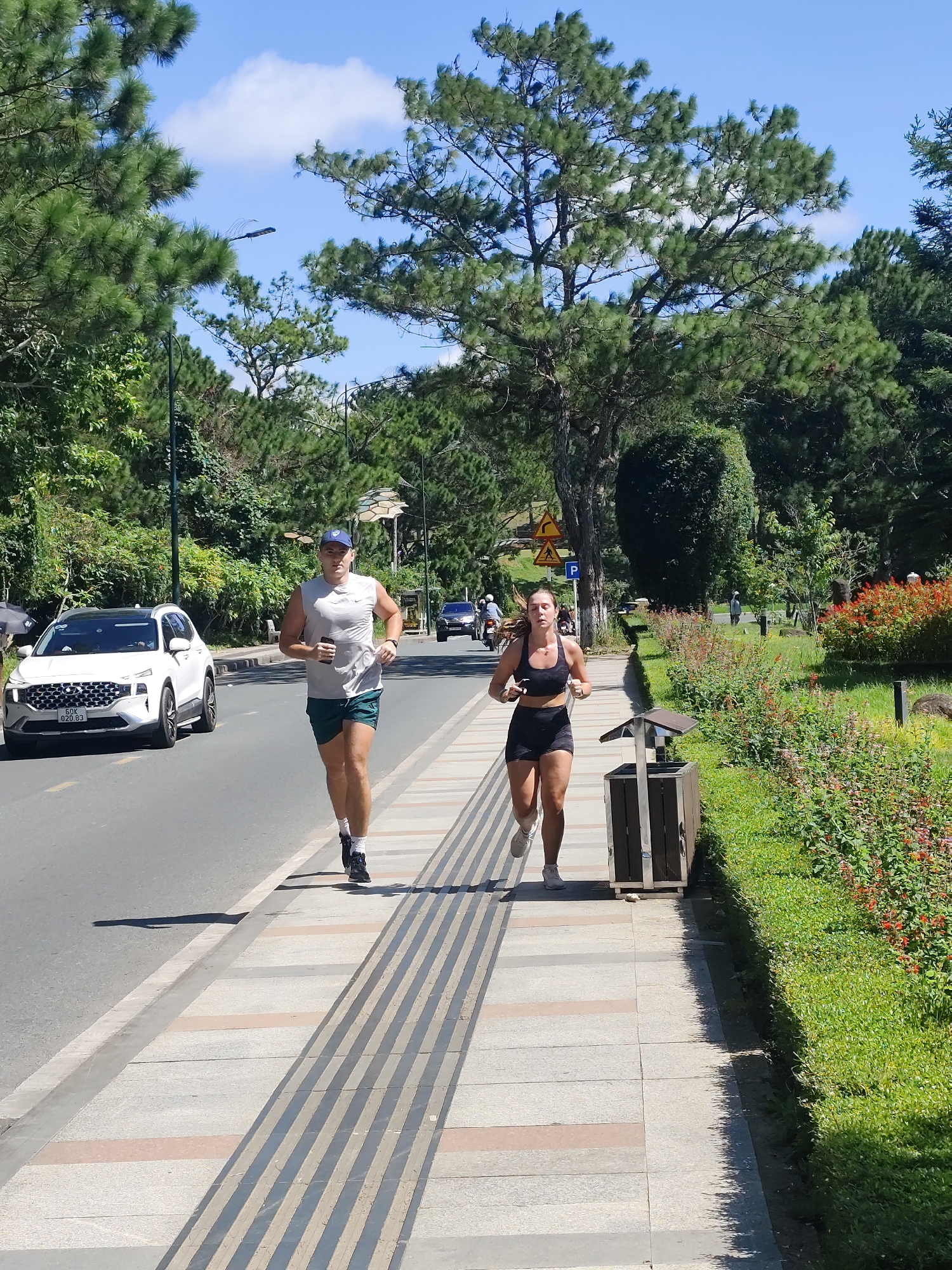 A man and a woman are jogging on a sidewalk next to a road. The man, wearing a white tank top and green shorts, is in the foreground, running towards the camera. The woman, in a black sports bra and black shorts, is slightly behind and to his right, also running towards the camera. They are on a sidewalk with textured paving stones. To their left is a road with a white SUV and other vehicles. To their right is a strip of grass and flowering bushes, and beyond that, more trees and greenery, suggesting a park-like setting. The background is filled with lush green trees and a clear blue sky. The scene appears to be in Xuân Hương - Đà Lạt, Vietnam, a location known for its scenic beauty and recreational areas, evidenced by the well-maintained sidewalk and roadside landscaping.