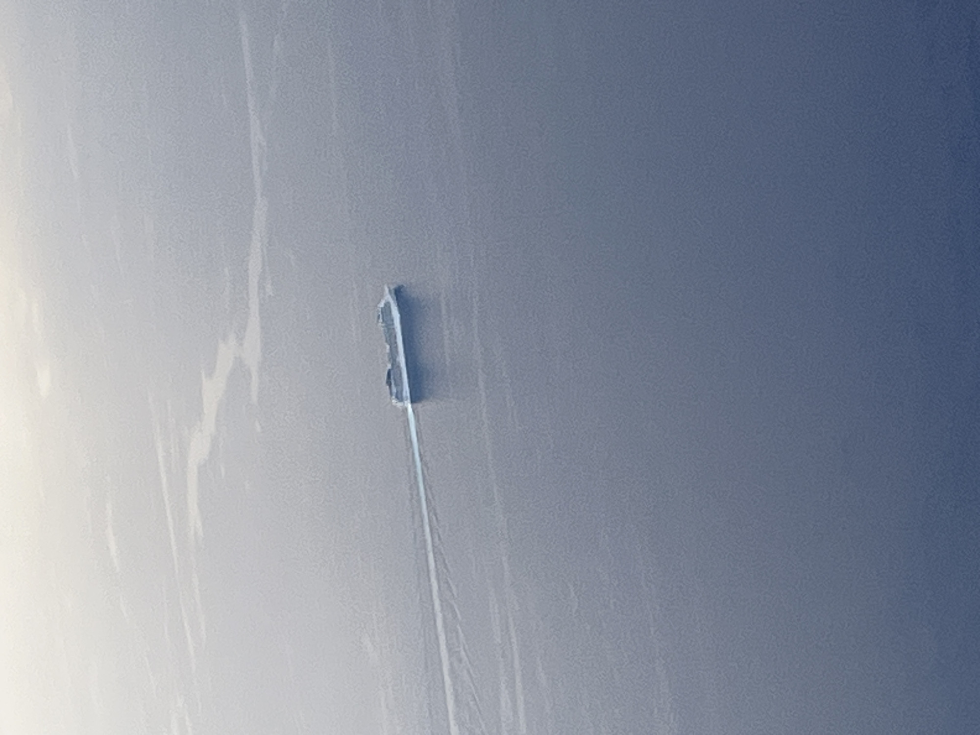 A distant aerial shot depicts a ship moving through a body of water. The water occupies the majority of the scene and appears relatively calm, with a blue-gray hue. The ship, positioned towards the center, has a dark silhouette. A trail extends from the back of the ship, indicating its movement direction. The sky or atmosphere in the background is a lighter shade of gray.