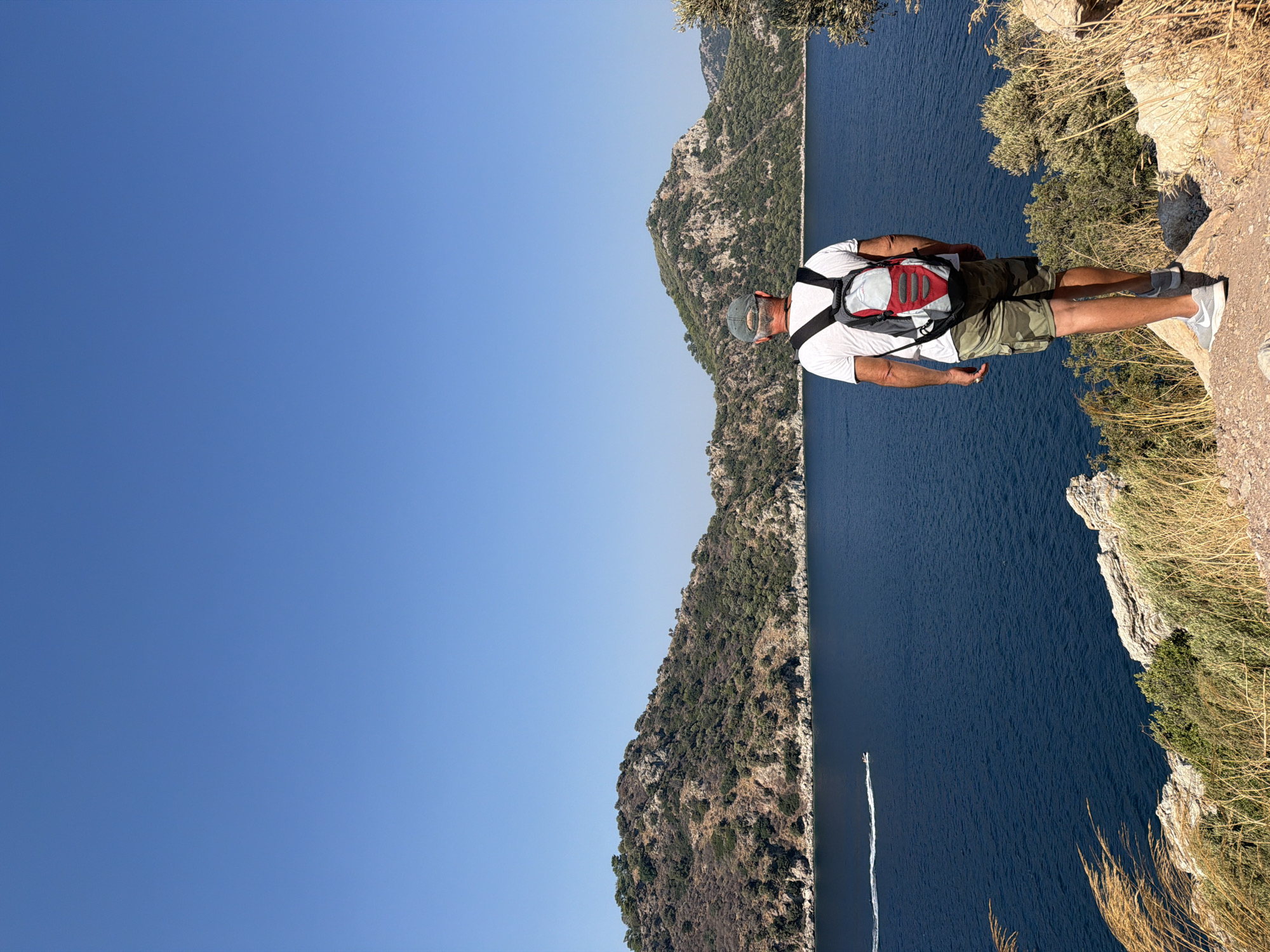 A man stands with his back to the camera on a rocky cliff overlooking a body of deep blue water. He is wearing a white t-shirt, camouflage shorts, a grey baseball cap, and a backpack with red and grey accents. The backpack is partially obscuring his head. His arms are relaxed at his sides. The water is calm and reflects the clear blue sky. In the distance, green, rocky hills rise from the water's edge, sparsely covered with vegetation. A small white boat with a wake is visible on the water, heading towards the left side of the frame. The foreground consists of dry grasses and rocks. The overall impression is a sunny day in a coastal, mountainous region, likely in or around Marmaris, Turkey.