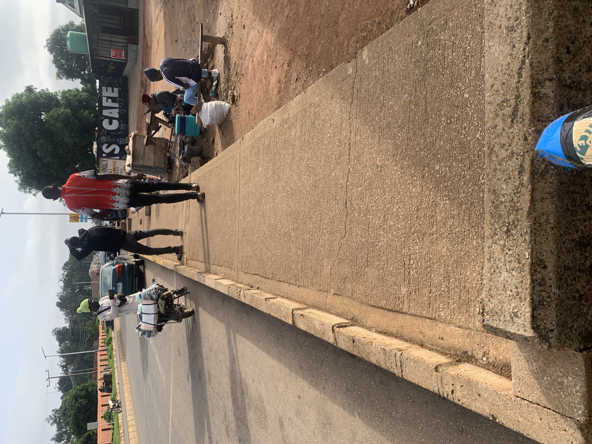 The image displays a lively street scene in Barakin Lerawi, Nigeria, under a bright, clear sky, indicating daytime. The perspective is looking down a sidewalk that runs parallel to a paved road.
On the sidewalk, a man in a distinctive red and white patterned shirt walks away from the camera, followed by another man in a black hoodie. Further down the street, a man wearing a light green hat is positioned beside a motorcycle heavily loaded with bundled textiles or other goods, and he appears to be talking on a mobile phone. A green sedan is parked on the street behind him. A small yellow sign reading "BUS STOP" is visible near the road.
Adjacent to the sidewalk, a dirt area hosts what appears to be makeshift stalls or vendors. Two individuals are crouching or sitting among various items, including a blue bucket, a white sack, and wooden structures, suggesting commercial activity. Behind these vendors, a prominent black sign with white, stylized letters clearly displays "S CAFE". Partially visible text below this sign includes "ARMACY", "PLAZA", and "FIELD". A green water tank can be seen on the roof of a building behind the cafe sign.
The street features a concrete curb separating the sidewalk from the asphalt road. Trees line parts of the street in the distance, and utility poles are also visible. On the far right edge of the frame, a blue and white object, possibly a part of a vehicle or equipment, is partially captured. The overall scene captures typical daily life and street commerce in an urban or semi-urban Nigerian setting.