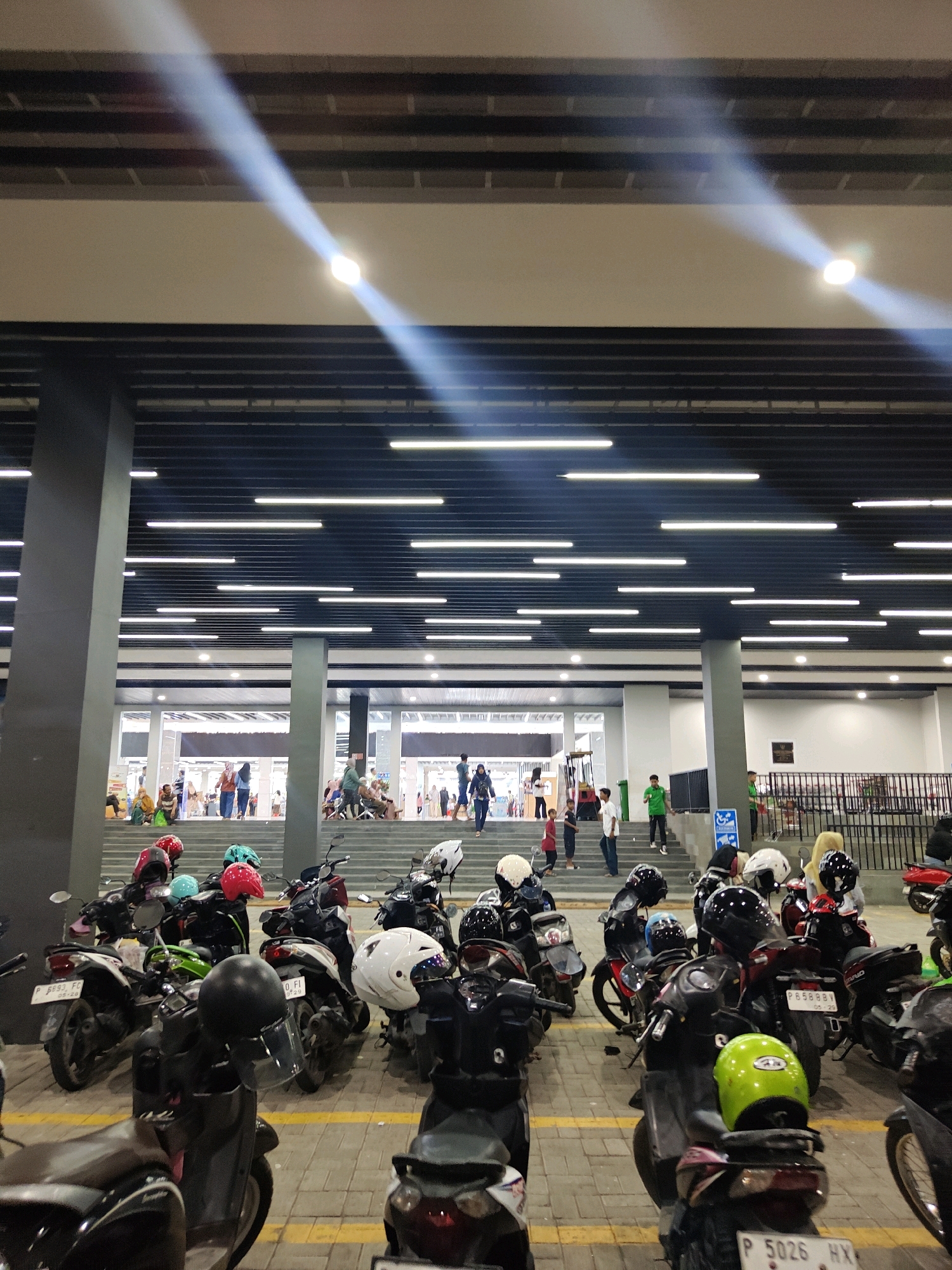 The image depicts an outdoor motorcycle parking area in front of a building, likely a shopping mall or entertainment venue, in Situbondo, Indonesia. The foreground is dominated by a row of parked motorcycles, many with helmets placed on them or their seats. The motorcycles are of various colors and models, suggesting a common mode of transportation in the area.
In the mid-ground, a set of wide stairs leads up to the building's entrance. People are seen ascending and descending the stairs, some walking individually and others in small groups. The attire of the people suggests a casual setting. Behind the stairs, the building's facade is visible, with large glass windows and entrances.
The background shows more of the building and its surroundings, with overhead lighting structures and what appears to be more parking or pedestrian areas. The overall lighting is a mix of natural light filtering from above and artificial lights from the building and lamps, creating some lens flare effects. The architectural style of the building, with its dark horizontal ceiling elements and prominent grey pillars, is modern. The ground is paved with interlocking brick tiles. The scene conveys a sense of everyday activity and public space.