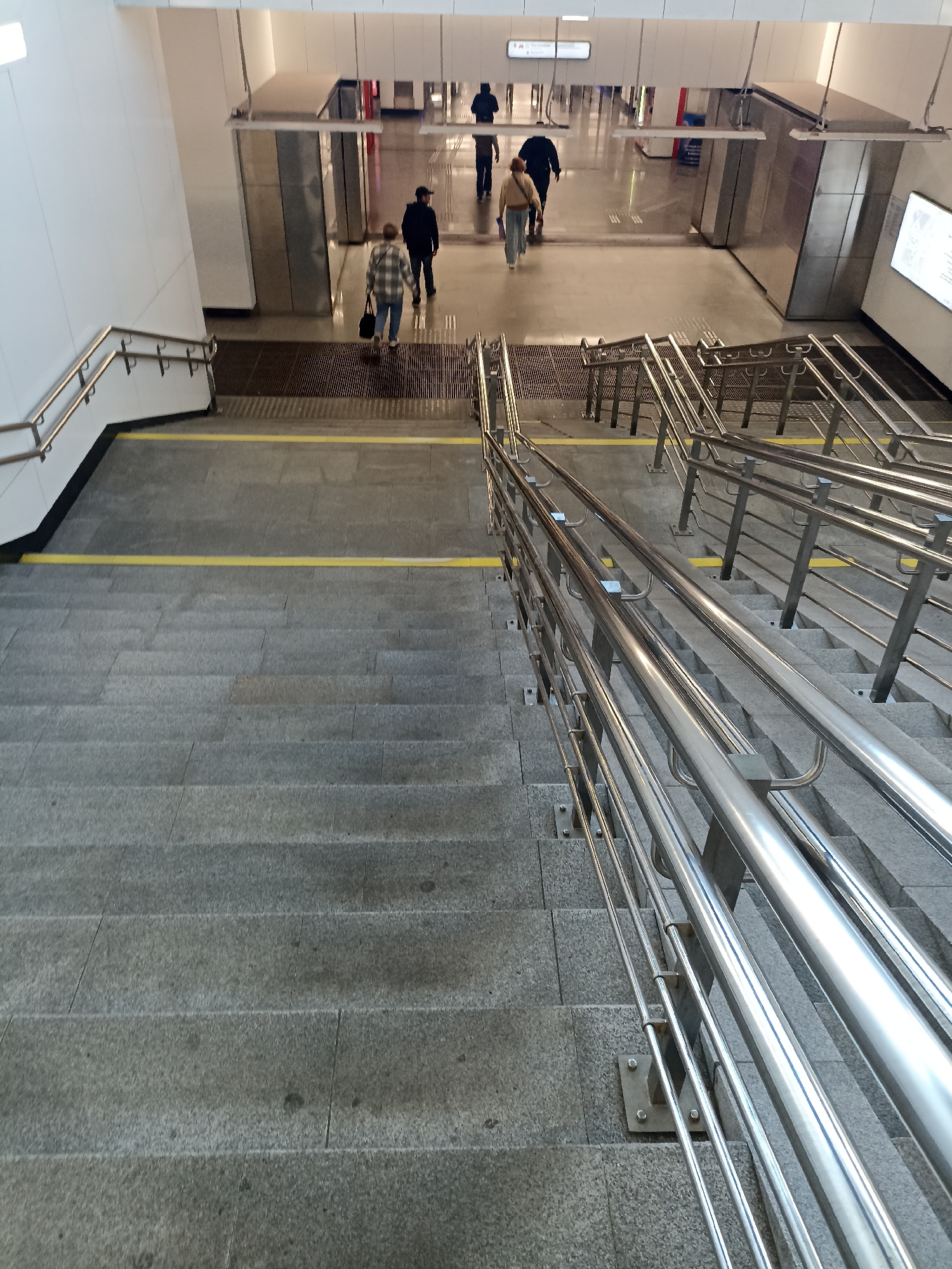 The image shows a view looking down a wide set of grey stone stairs with polished metal handrails in an indoor public space, likely a subway station or a large building. Two yellow lines are visible on the stair landing. Several people are visible in the background at the top of the stairs, walking away from the camera. One person in the foreground is walking down the stairs, also away from the camera, carrying a black bag. The environment is clean and modern, with white walls and metal accents.